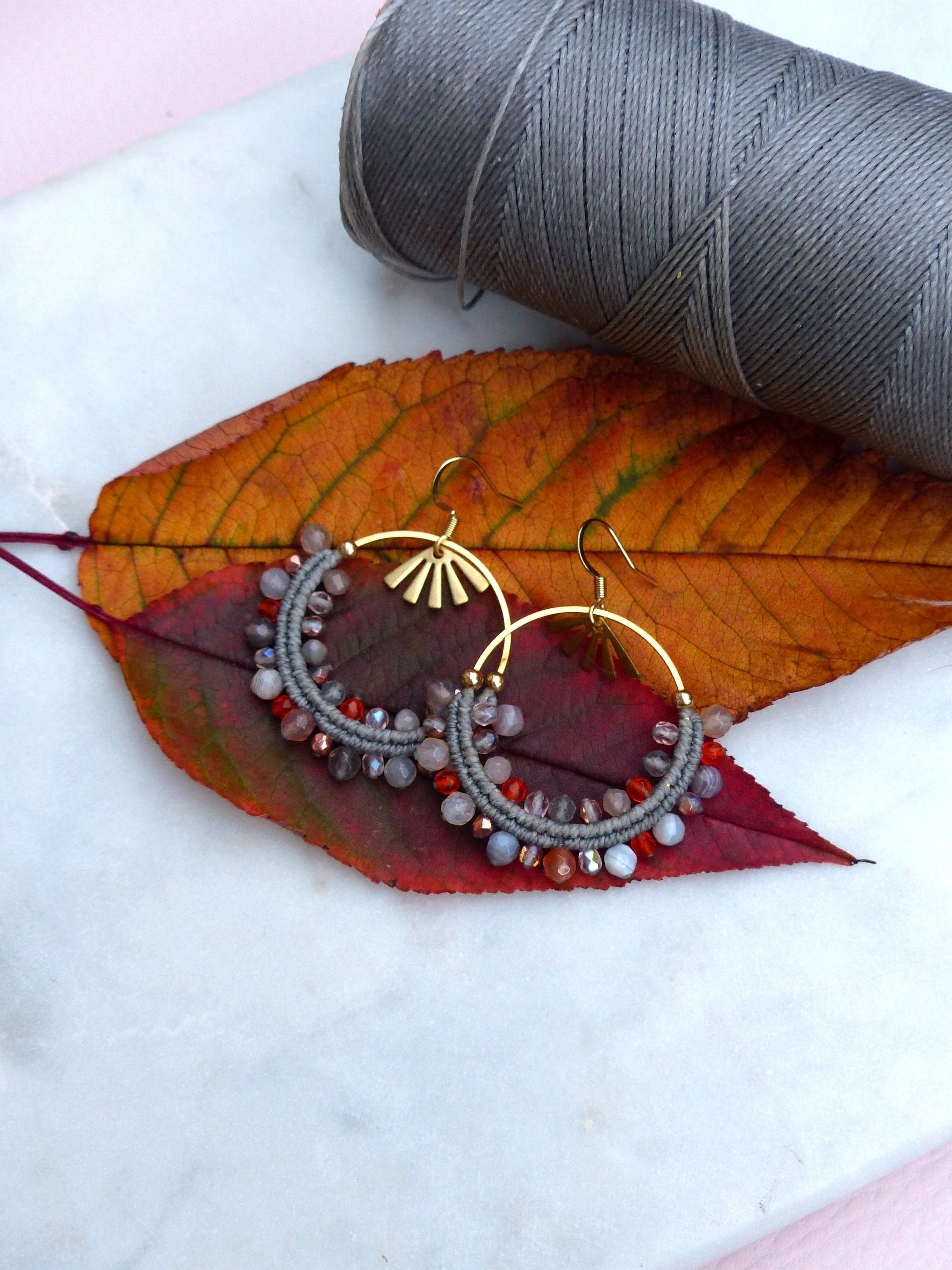 Peut inclure: Une paire de boucles d'oreilles créoles dorées avec des perles grises et rouges. Les boucles d'oreilles sont sur une surface blanche avec une feuille d'automne.