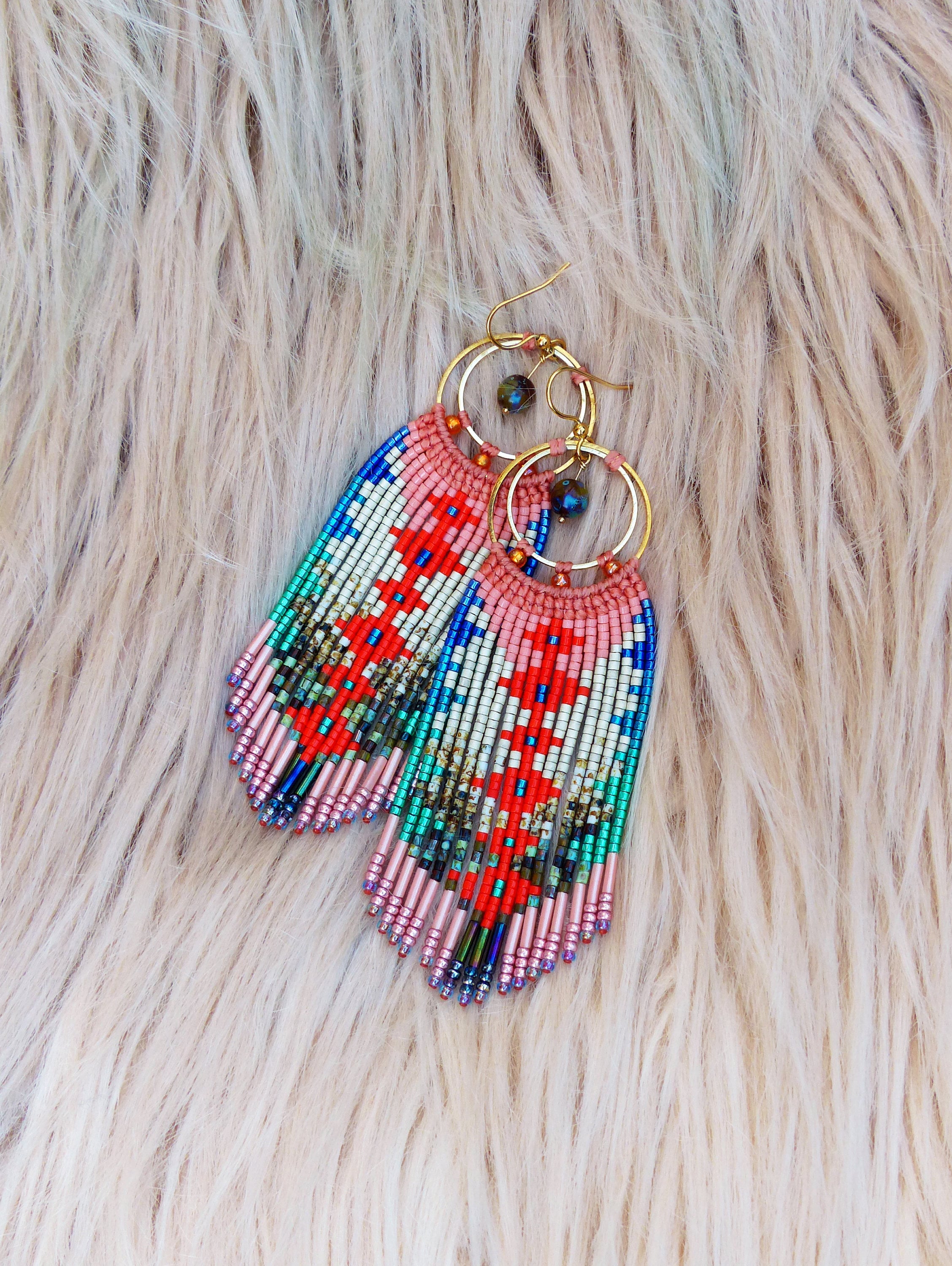 Peut inclure: Une paire de longues boucles d'oreilles en perles avec un motif géométrique dans des tons de rouge, rose, bleu et vert. Les boucles d'oreilles sont suspendues à des cercles dorés avec un accent de pierre précieuse bleu foncé.