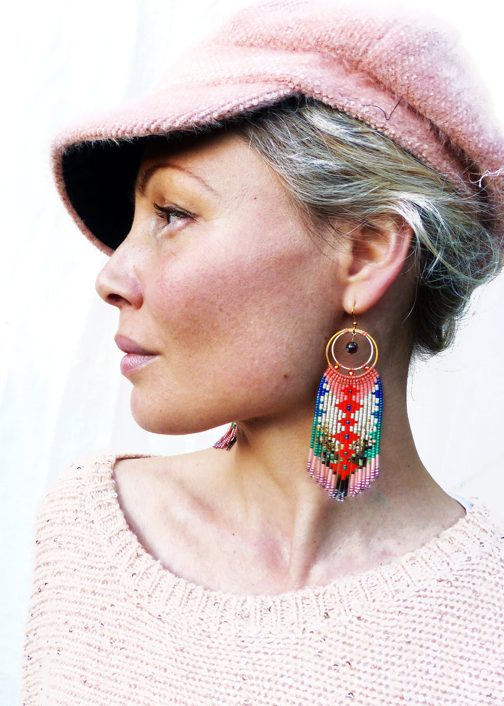 Peut inclure: Une femme portant un chapeau en tricot rose et un pull rose avec de longues boucles d'oreilles en perles colorées. Les boucles d'oreilles ont un motif géométrique et un anneau en or.