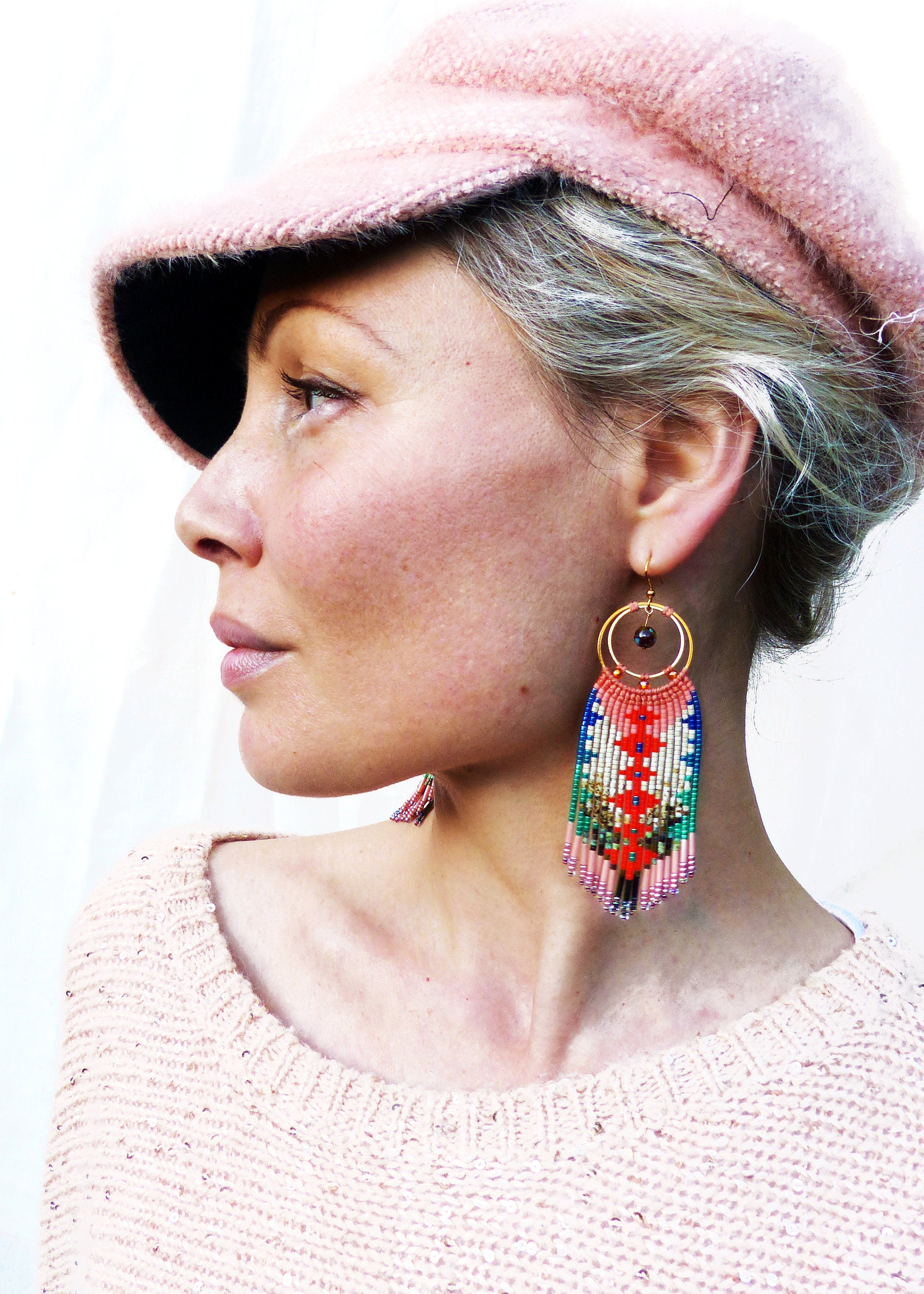 Peut inclure: Une femme portant un chapeau en tricot rose et un pull rose avec de longues boucles d'oreilles en perles colorées. Les boucles d'oreilles ont un motif géométrique et un anneau en or.
