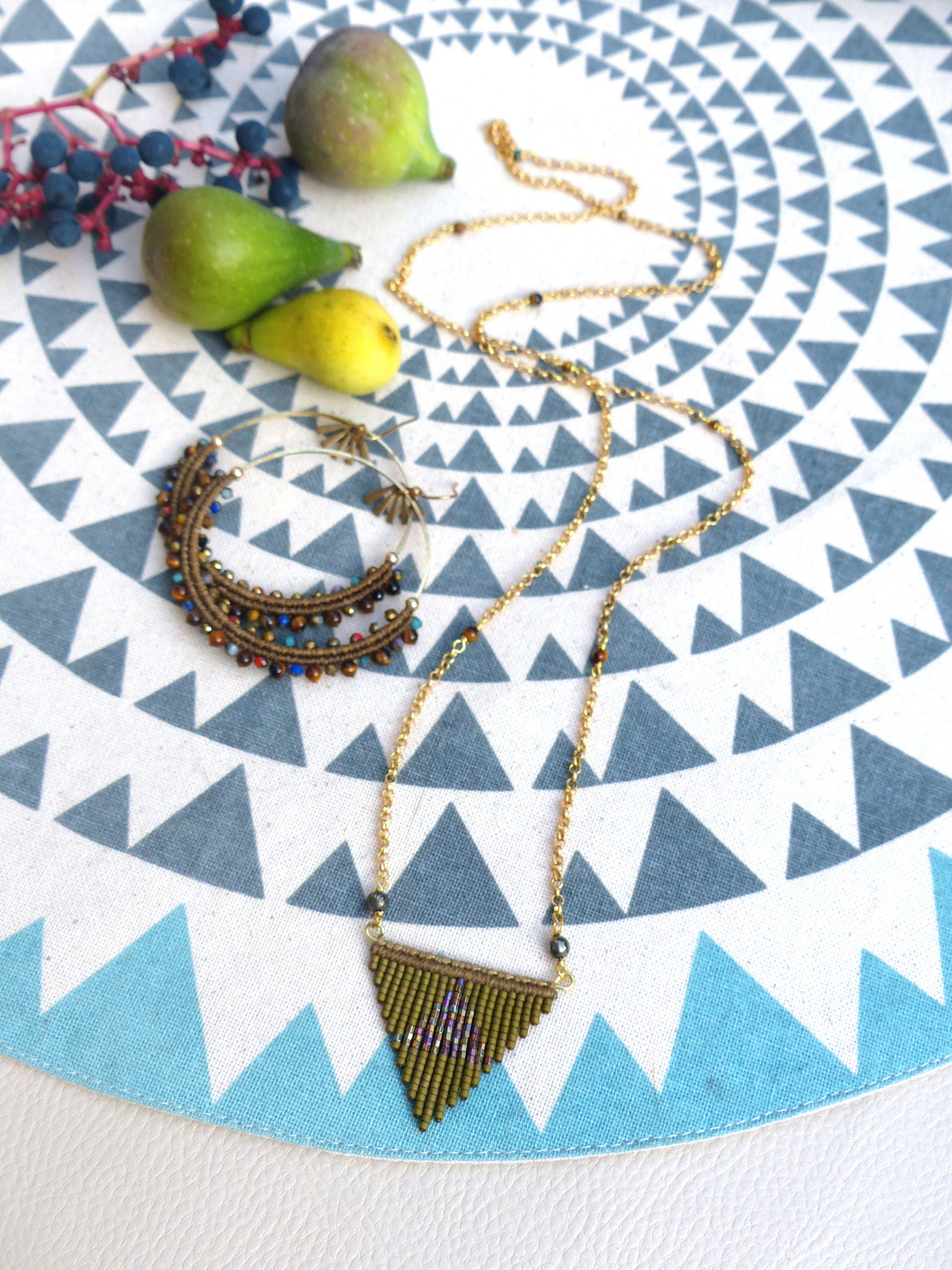 Peut inclure: Un collier en chaîne dorée avec un pendentif triangulaire en perles brunes. Le pendentif est un motif géométrique avec une frange de perles. Le collier est sur un fond blanc avec un motif bleu et blanc.