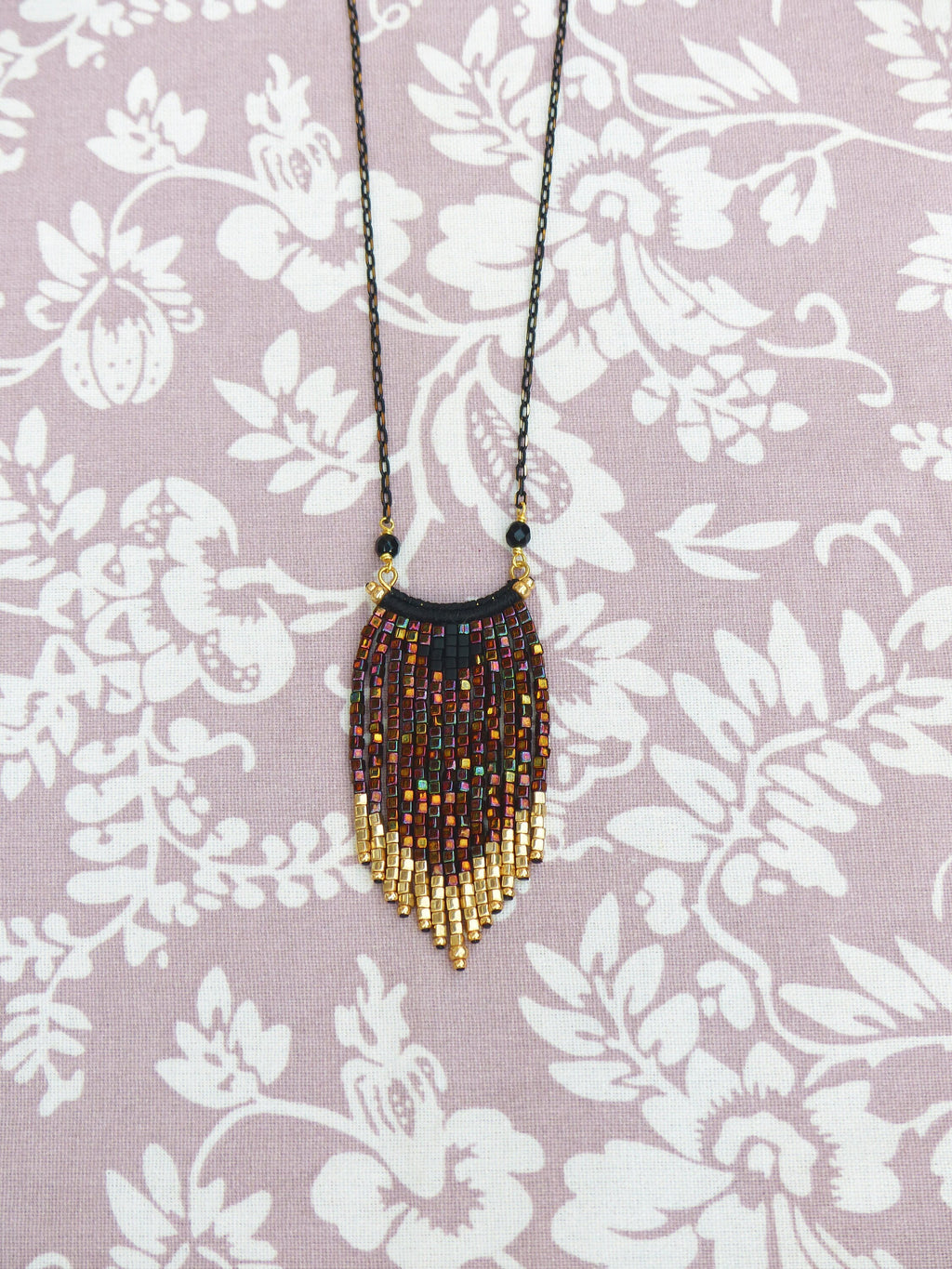 Peut inclure: Un collier de perles avec une chaîne noire et un pendentif en frange. Le pendentif est fait de perles dorées et brunes et a une forme géométrique.
