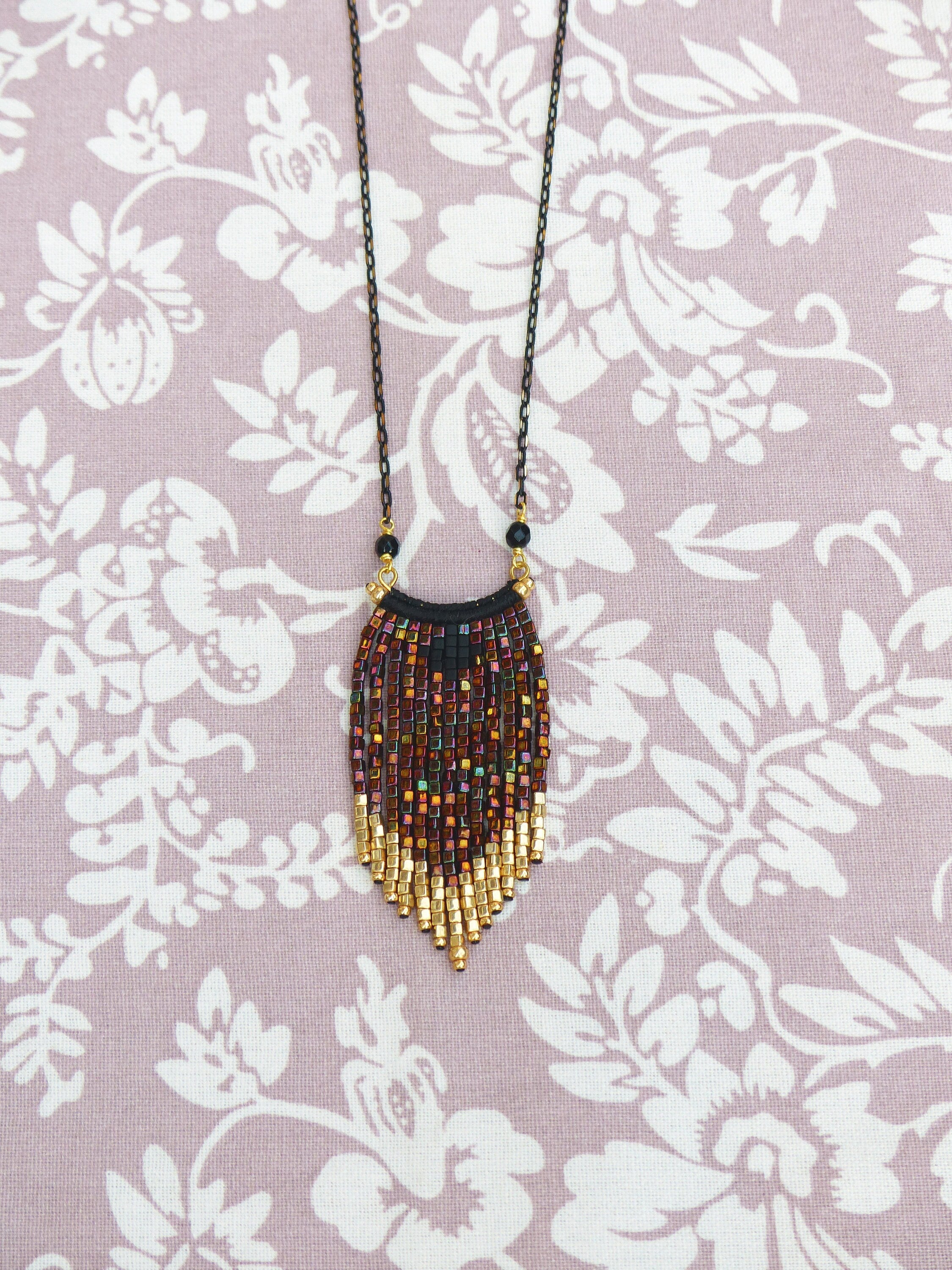 Peut inclure: Un collier de perles avec une chaîne noire et un pendentif en frange. Le pendentif est fait de perles dorées et brunes et a une forme géométrique.