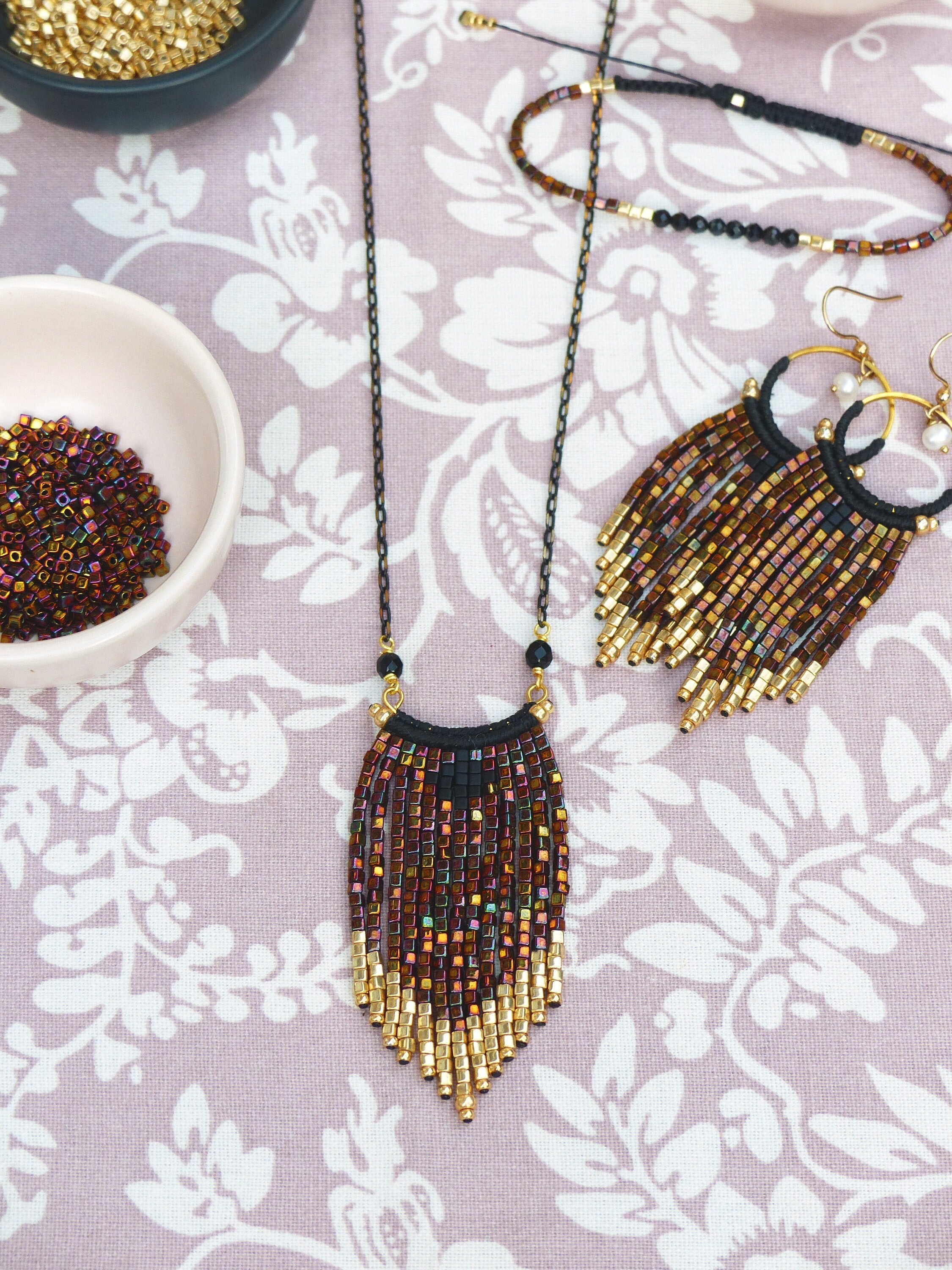 Peut inclure: Un collier de perles dorées et brunes avec un pendentif en frange. Le collier est composé de petites perles carrées et possède un cordon noir en haut. Le collier est présenté sur un fond de tissu floral rose.