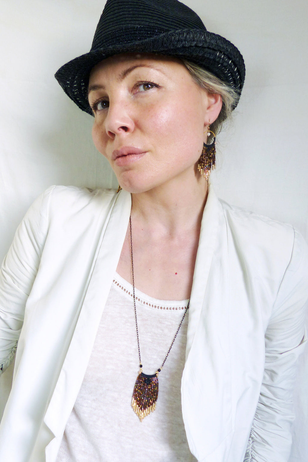 Peut inclure: Une femme portant un blazer blanc, un fedora noir et un collier et des boucles d'oreilles en perles dorées et brunes. Le collier et les boucles d'oreilles sont tous deux de style frange.
