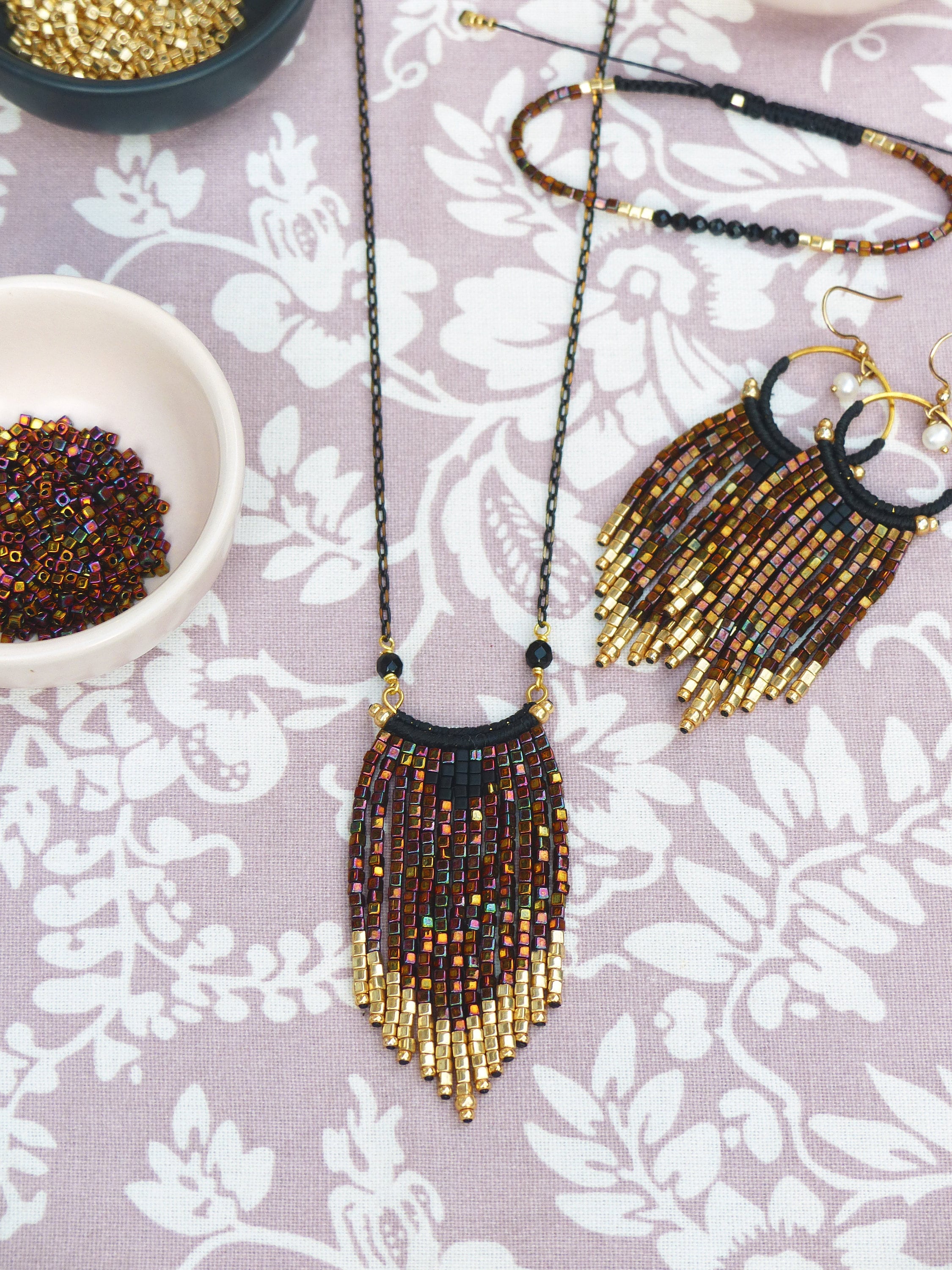 Peut inclure: Un collier de perles dorées et brunes avec un pendentif en frange. Le collier est composé de petites perles carrées et possède un cordon noir en haut. Le collier est présenté sur un tissu floral rose et blanc.