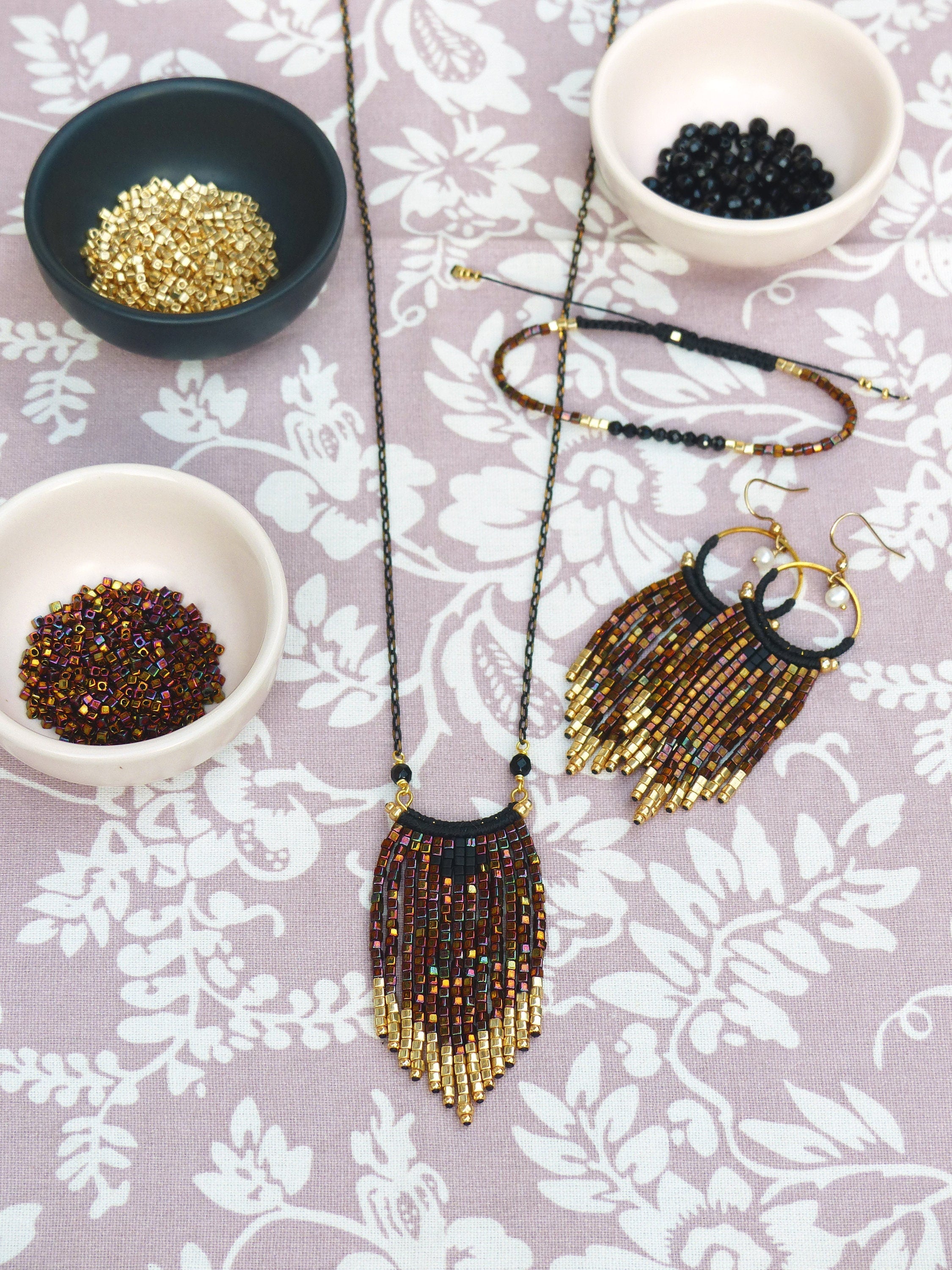 Peut inclure: Un collier de perles dorées et brunes avec un design en frange. Le collier est sur une table avec des bols de perles de différentes couleurs.