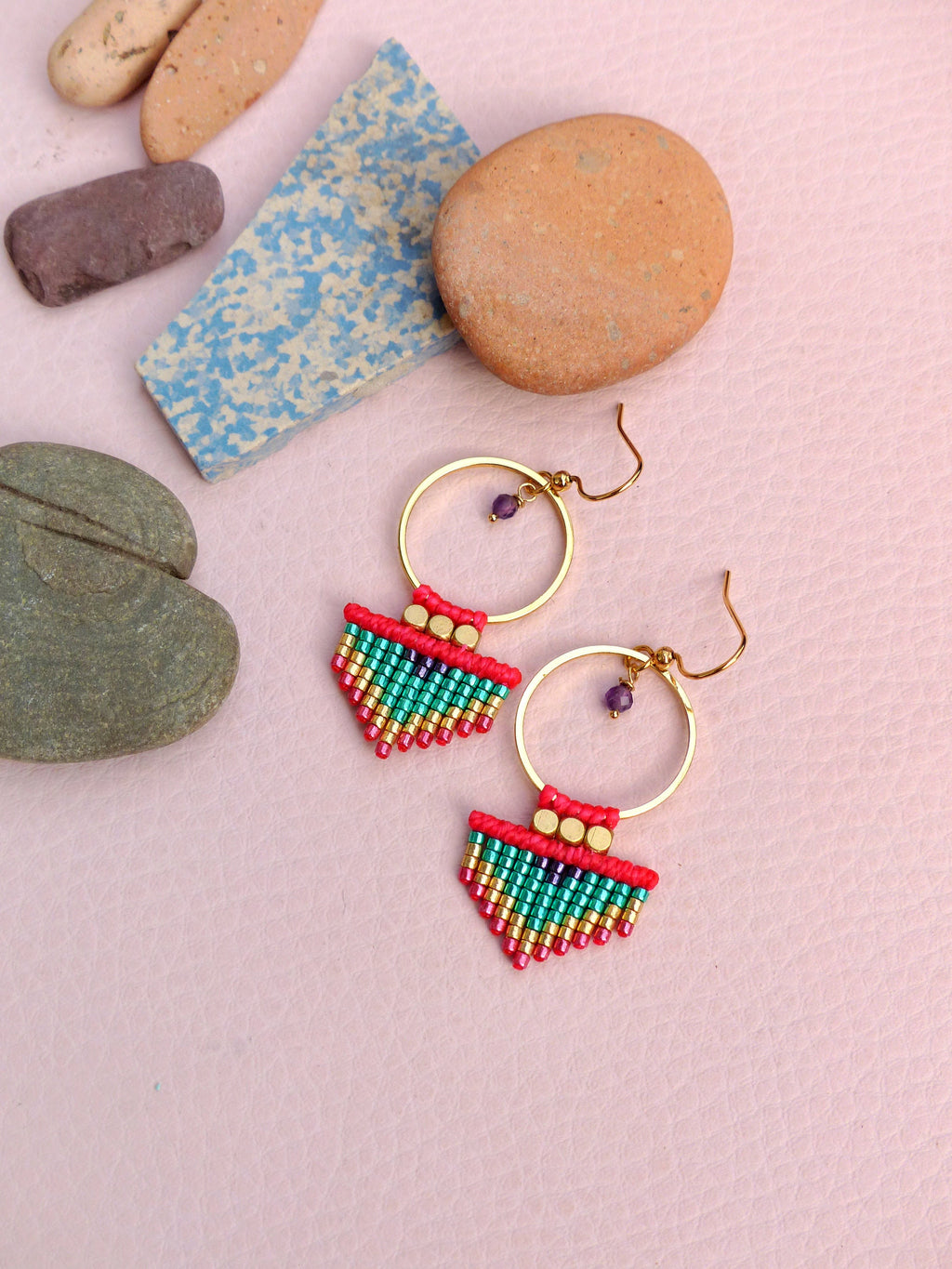 Peut inclure: Une paire de boucles d'oreilles créoles dorées avec une frange de perles. La frange est composée de perles rouges, vertes et dorées. Les boucles d'oreilles ont une petite perle violette suspendue au cerceau.