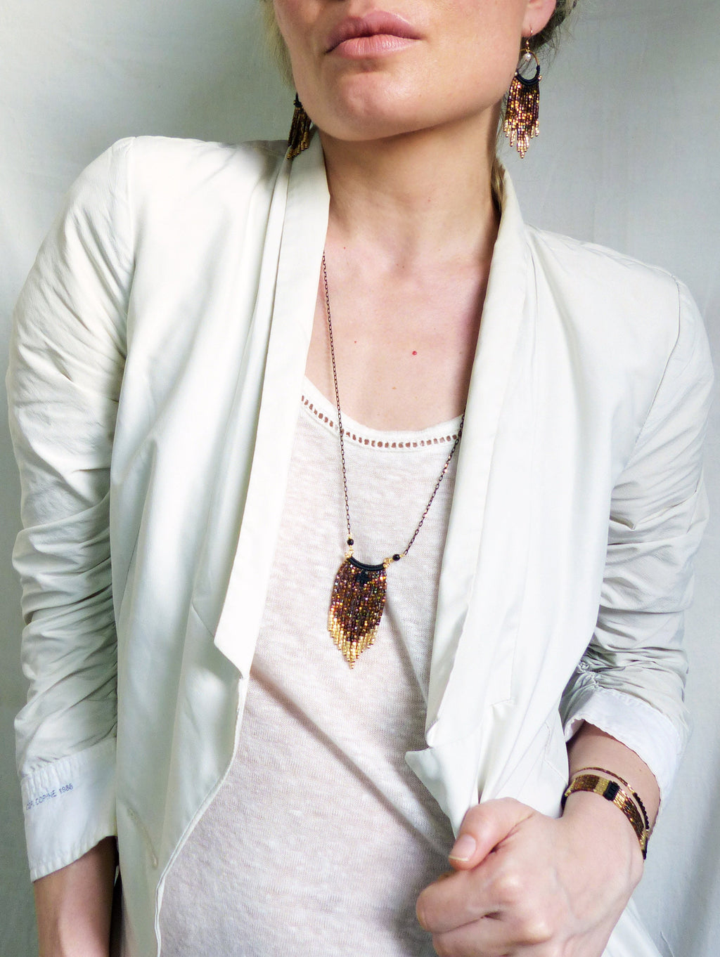 Peut inclure: Un blazer blanc avec un collier et des boucles d'oreilles assortis en perles brunes et dorées. Le collier présente un pendentif en forme de larme.