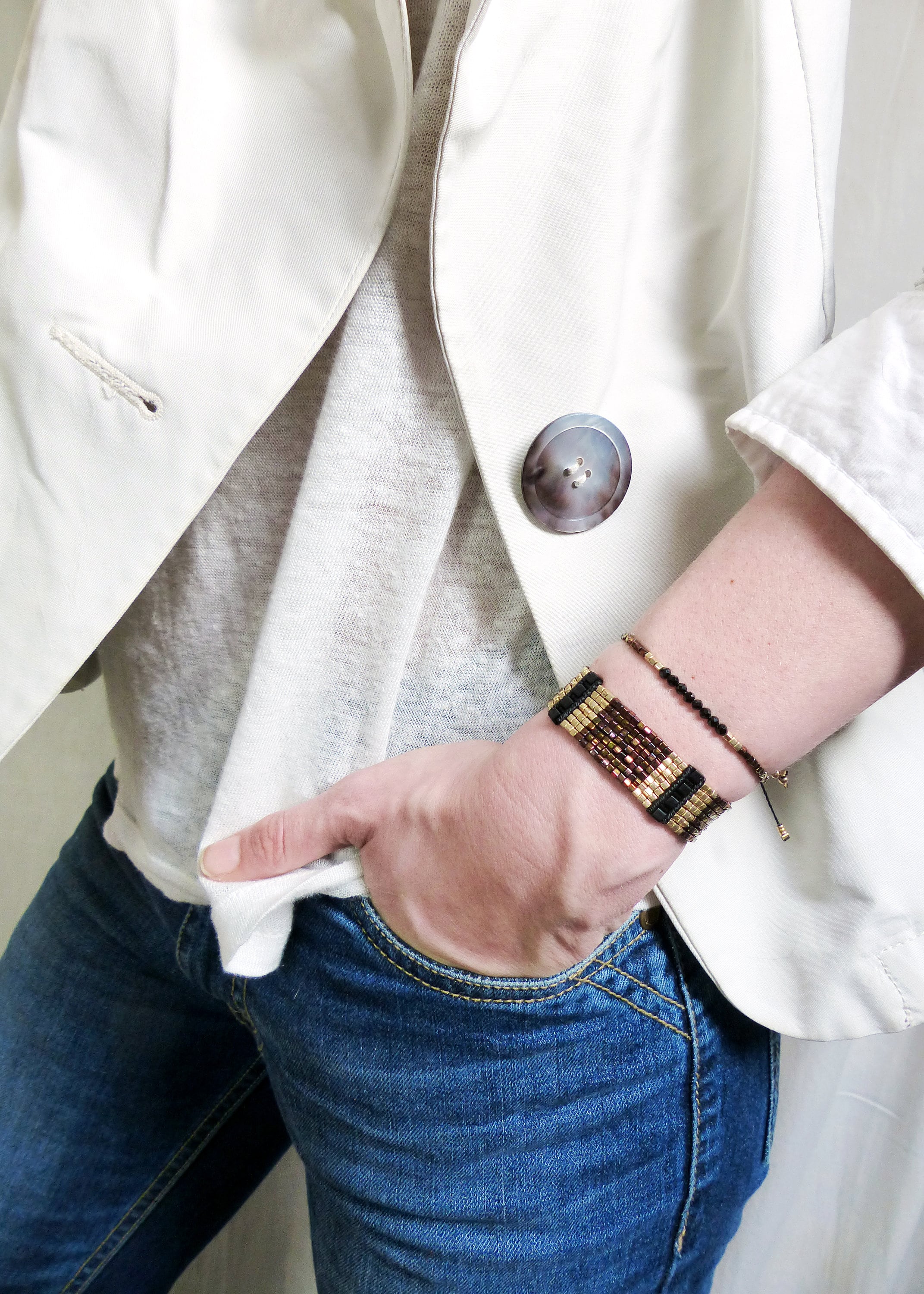 Peut inclure: Une personne portant un blazer blanc, un t-shirt blanc et un jean bleu. Elle porte deux bracelets au poignet, l'un est un bracelet en perles avec des perles brunes, noires et dorées, et l'autre est un bracelet fin en perles noires.