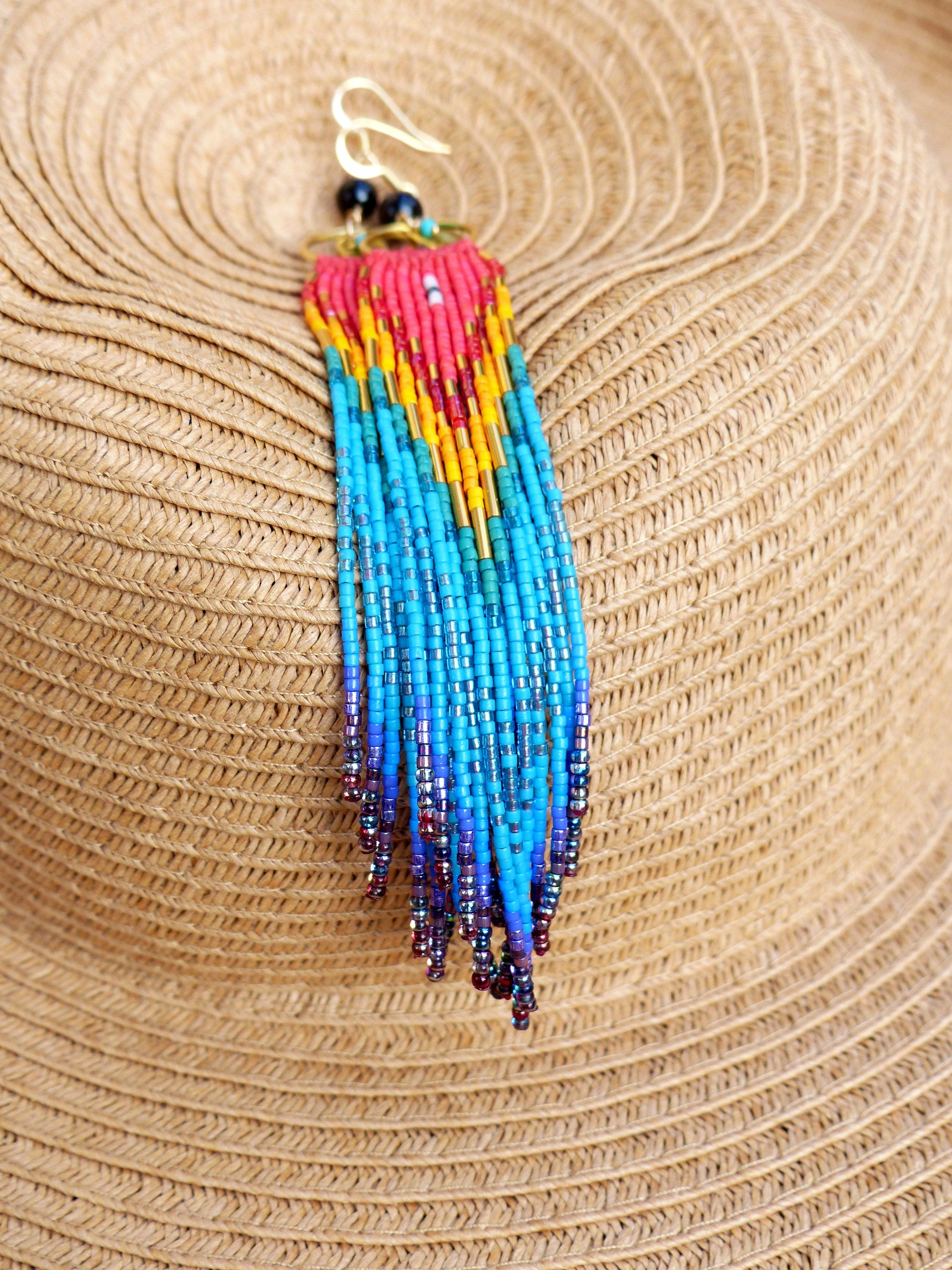 Peut inclure: Une paire de longues boucles d'oreilles en perles colorées avec un dégradé arc-en-ciel de perles rouges, orange, jaunes, vertes, bleues et violettes. Les boucles d'oreilles sont suspendues à des crochets dorés.