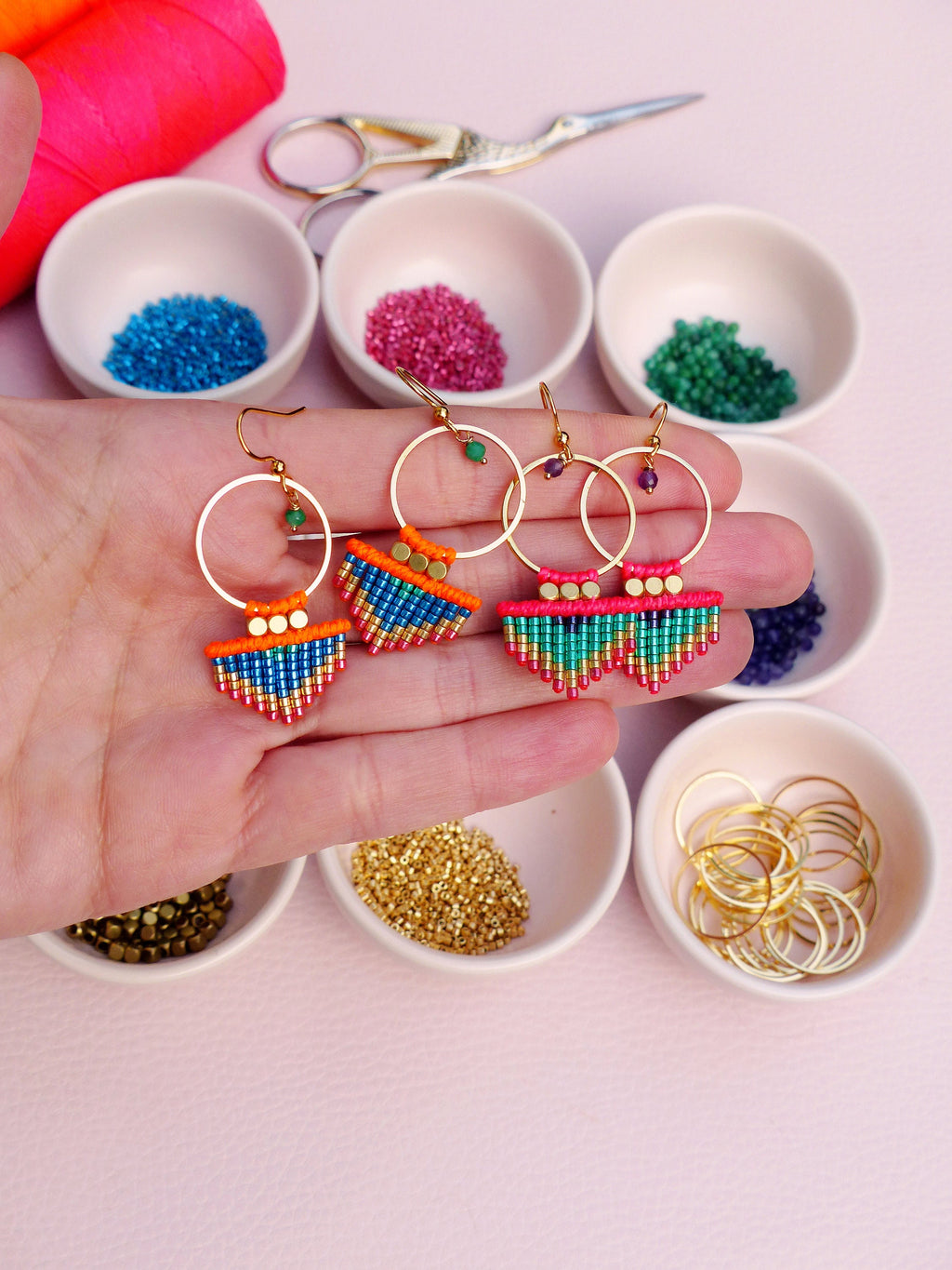 Peut inclure: Trois paires de boucles d'oreilles créoles dorées avec des franges de perles colorées. Les boucles d'oreilles sont présentées sur une main sur un fond rose. Les perles sont dans des tons de bleu, vert, orange et rose.