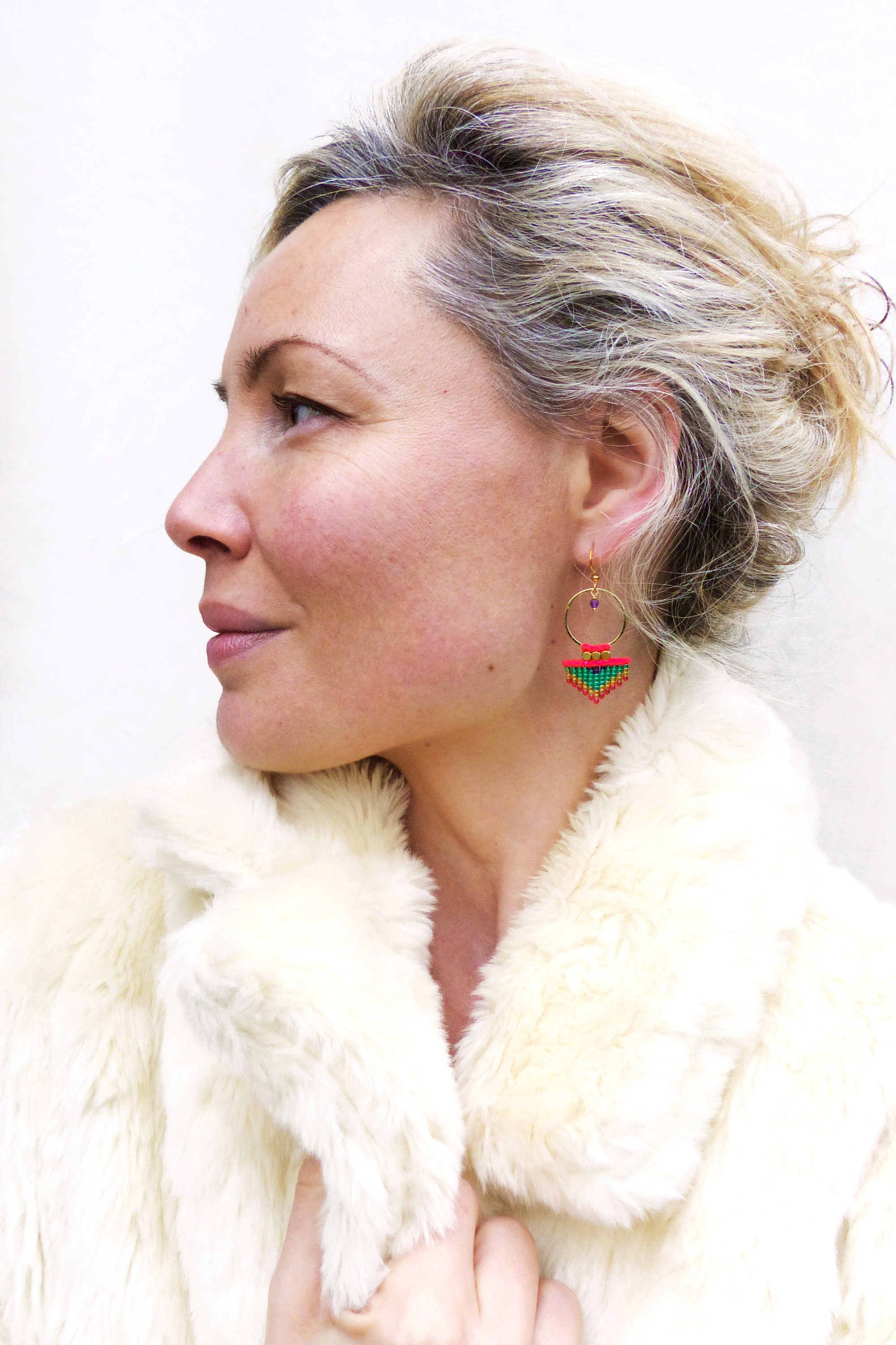 Peut inclure: Une femme portant une veste en fausse fourrure blanche avec une paire de créoles dorées avec un pendentif triangulaire en perles colorées.