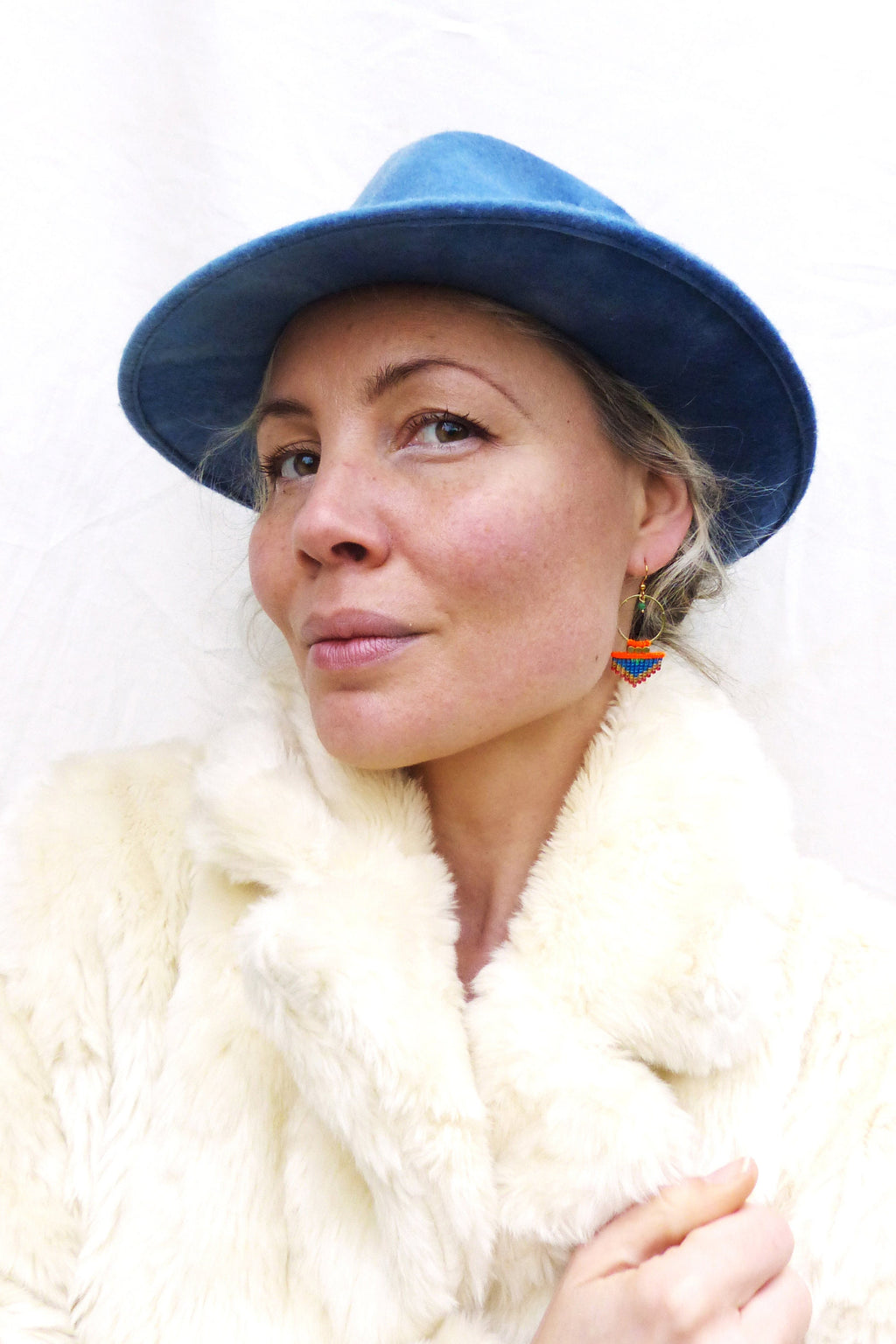 Peut inclure: Une femme portant un chapeau bleu et un manteau en fausse fourrure blanc. Elle porte des boucles d'oreilles en perles avec des accents orange, bleu et vert.