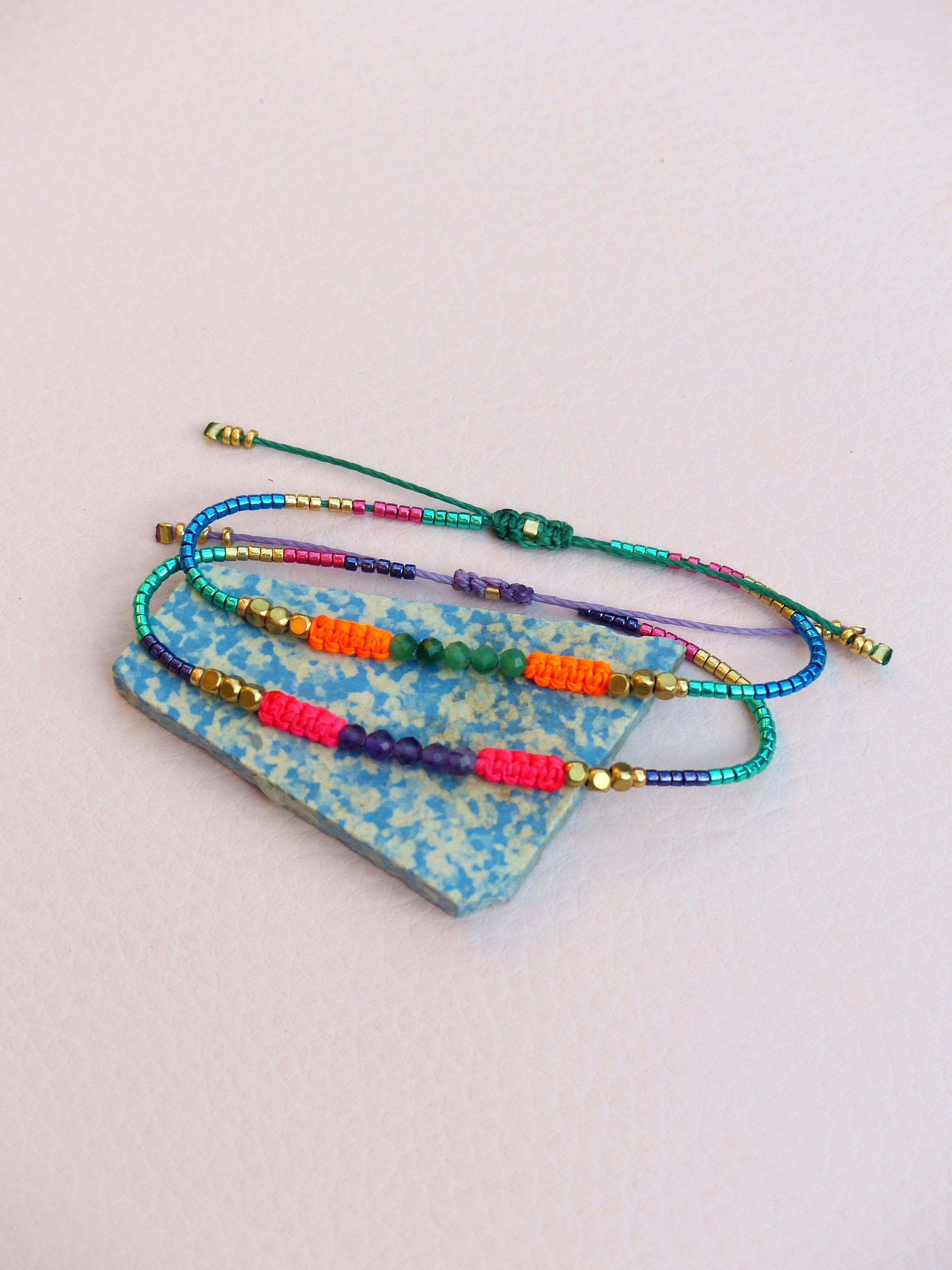 Peut inclure: Trois bracelets en perles colorées avec un mélange de perles vertes, violettes, orange et roses. Les bracelets sont liés ensemble et reposent sur une tuile à motifs bleus et blancs.