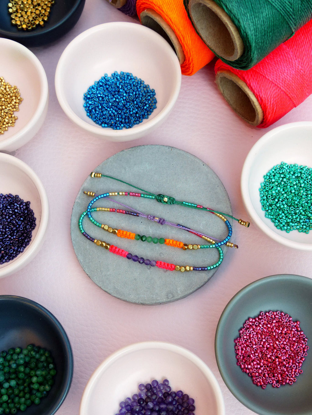 Peut inclure: Un assortiment de perles colorées dans de petits bols, dont de l'or, du bleu, du vert, du violet et du rose. Un bracelet fait avec ces perles est présenté sur un sous-verre en pierre grise.