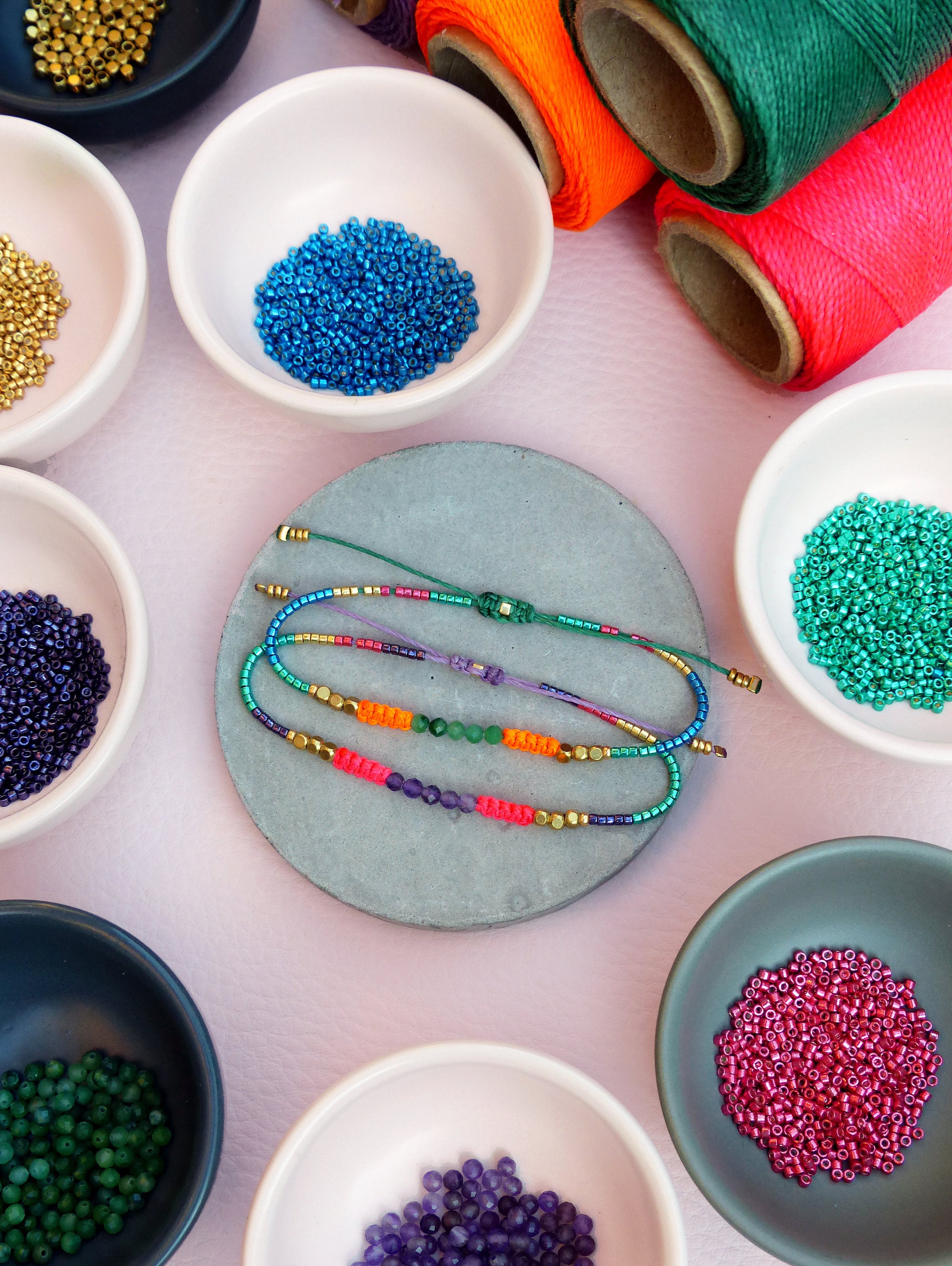Peut inclure: Un assortiment de perles colorées dans de petits bols, dont de l'or, du bleu, du vert, du violet et du rose. Un bracelet fait avec ces perles est présenté sur un sous-verre en pierre grise.