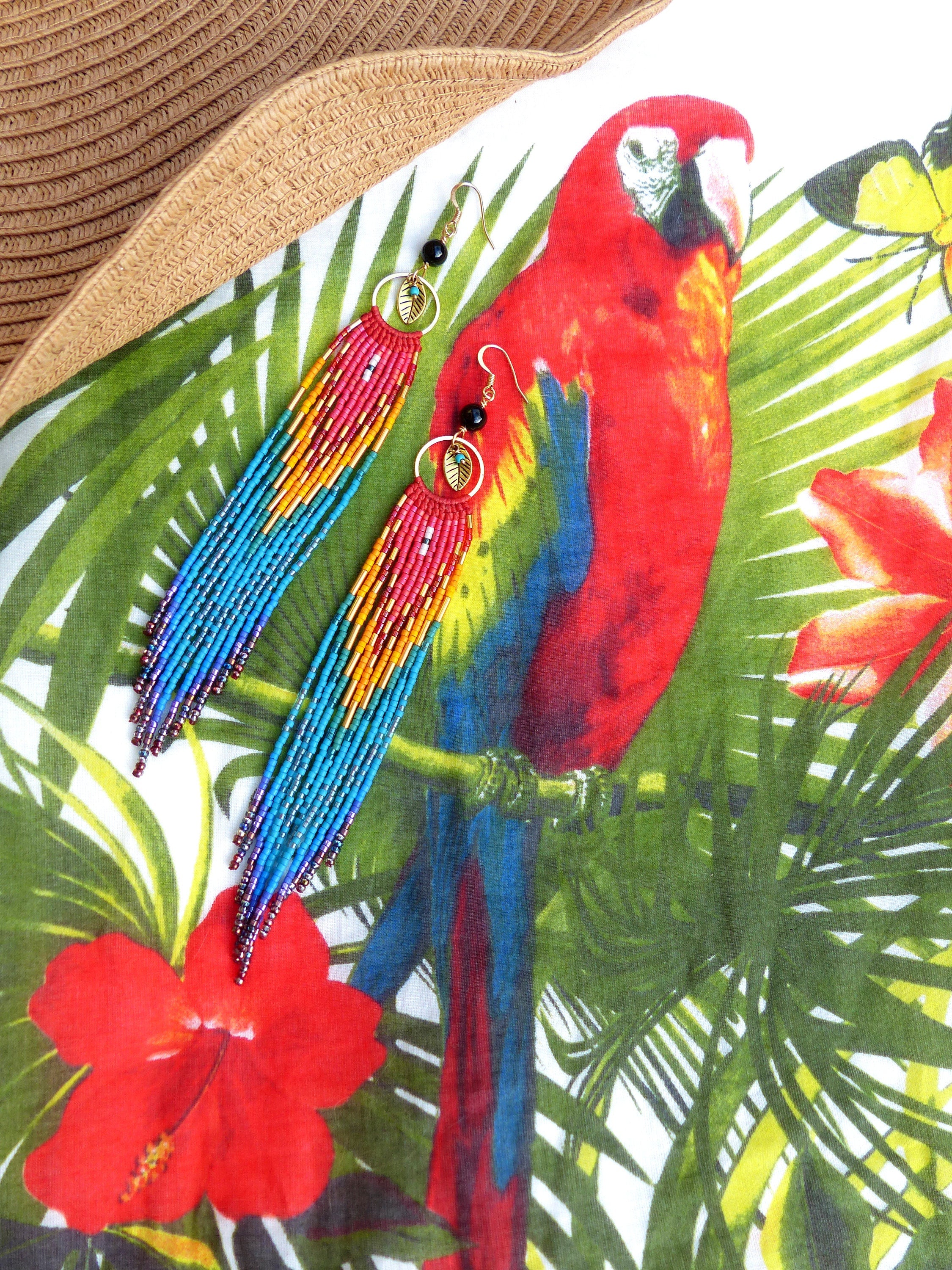 Peut inclure: Une paire de longues boucles d'oreilles en perles colorées avec des accents de feuilles d'or. Les boucles d'oreilles sont suspendues à un tissu à imprimé tropical avec un perroquet rouge.