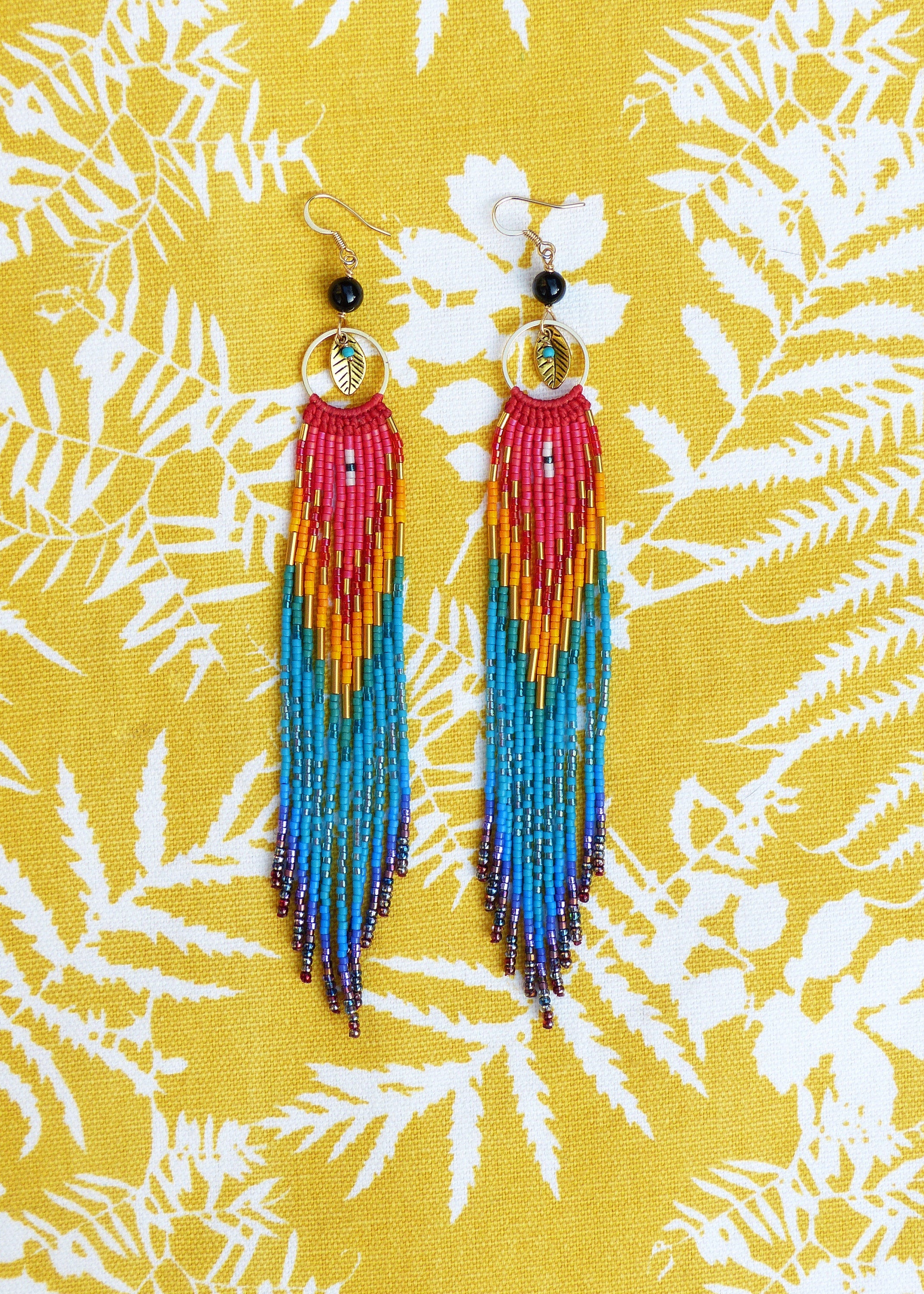 Peut inclure: Une paire de longues boucles d'oreilles en perles colorées avec un charme en feuille d'or. Les boucles d'oreilles sont faites de perles rouges, orange, jaunes, bleues et violettes.