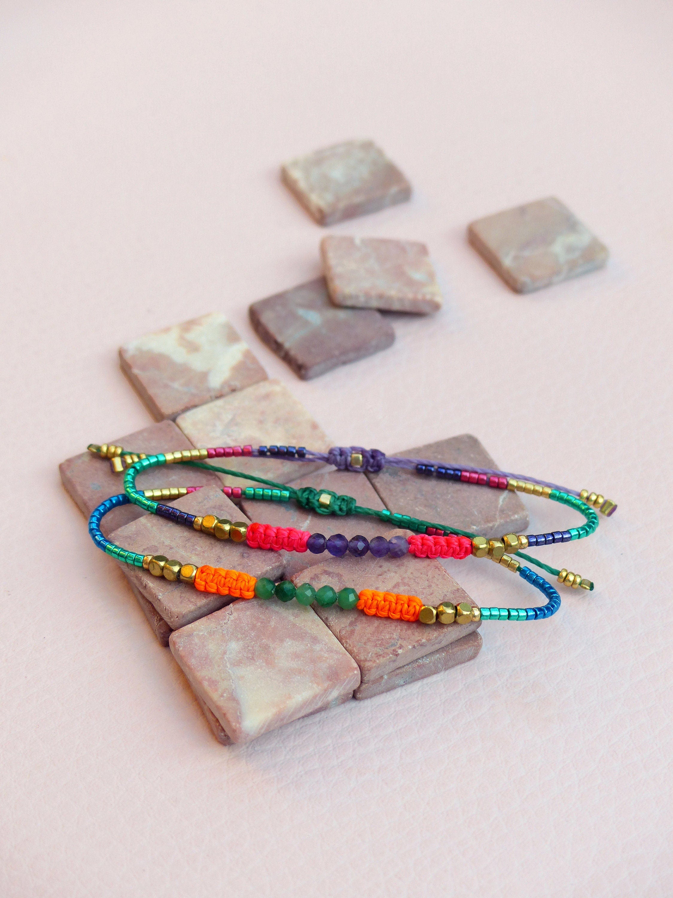 Peut inclure: Trois bracelets d'amitié colorés avec des perles et de petits accents dorés. Les bracelets sont superposés et se trouvent sur une surface rose.