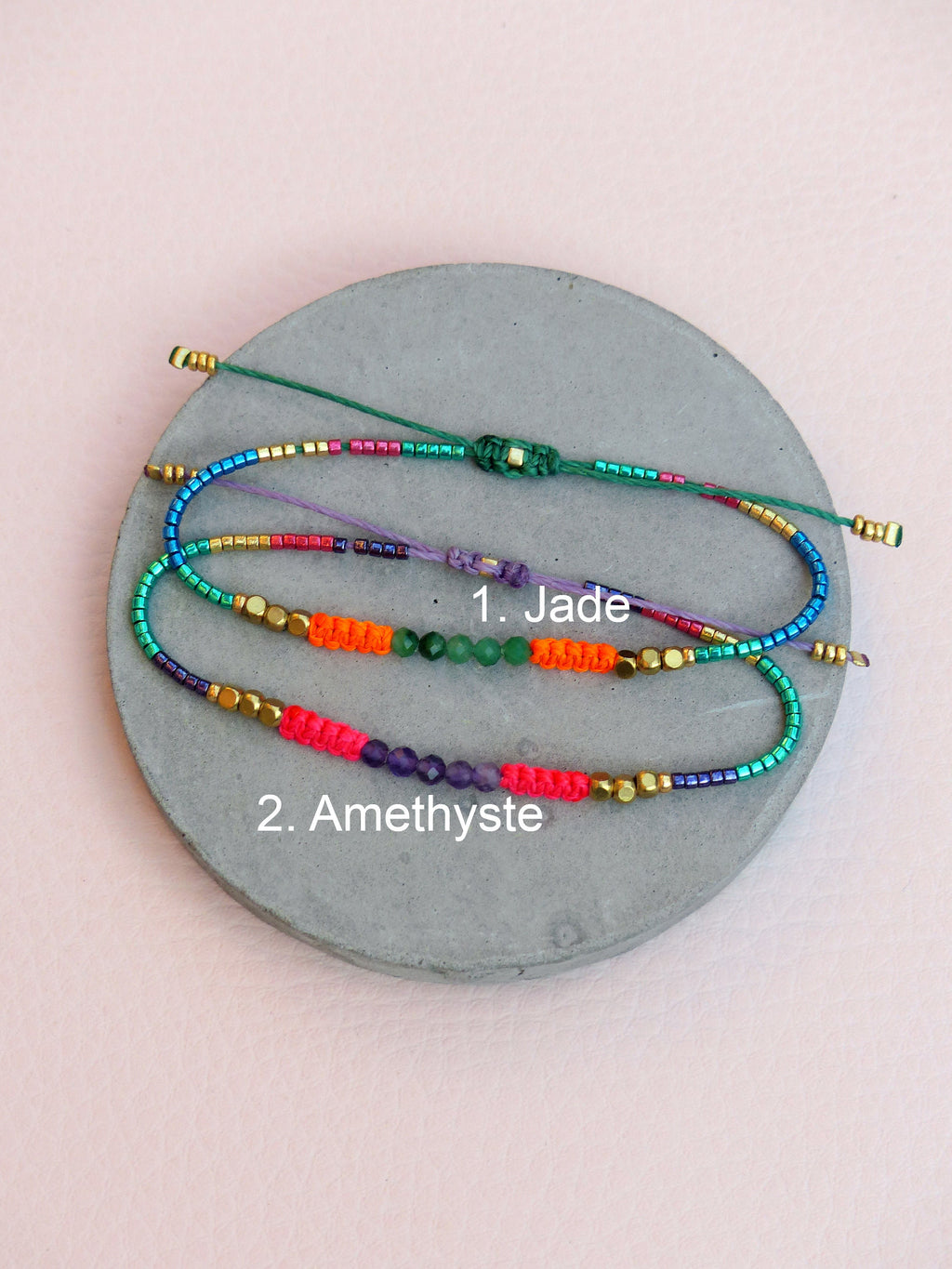 Peut inclure: Trois bracelets en perles colorées avec des perles de différentes couleurs et un charme en pierre précieuse. Le bracelet du haut a un charme en jade, le bracelet du milieu a un charme vert et le bracelet du bas a un charme en améthyste. Les bracelets sont sur une surface circulaire grise.