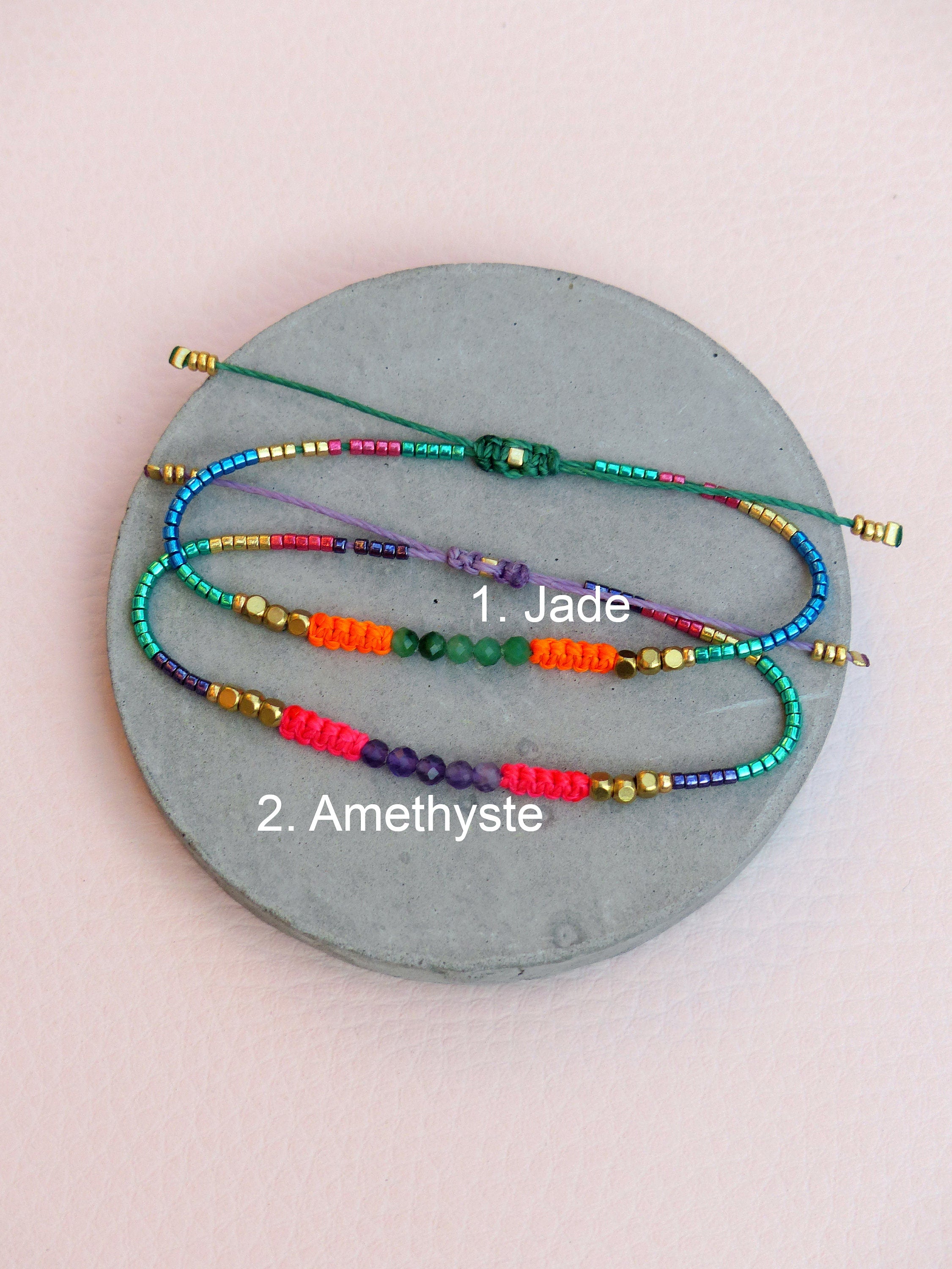 Peut inclure: Trois bracelets en perles colorées avec des perles de différentes couleurs et un charme en pierre précieuse. Le bracelet du haut a un charme en jade, le bracelet du milieu a un charme vert et le bracelet du bas a un charme en améthyste. Les bracelets sont sur une surface circulaire grise.