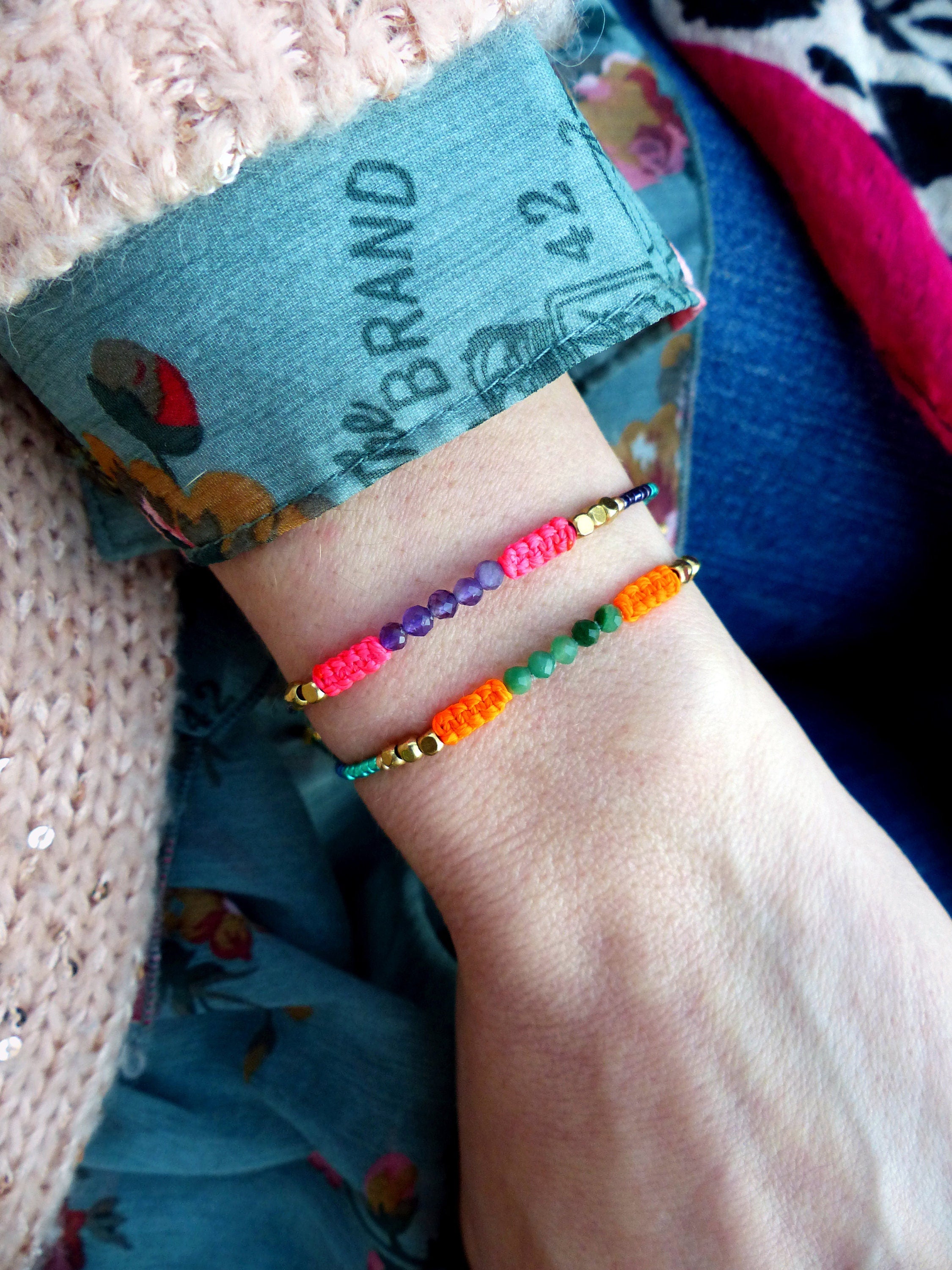 Peut inclure: Deux bracelets en perles colorées sur un poignet. Le bracelet du haut a des perles violettes, orange et roses avec des accents dorés. Le bracelet du bas a des perles vertes et orange avec des accents dorés.