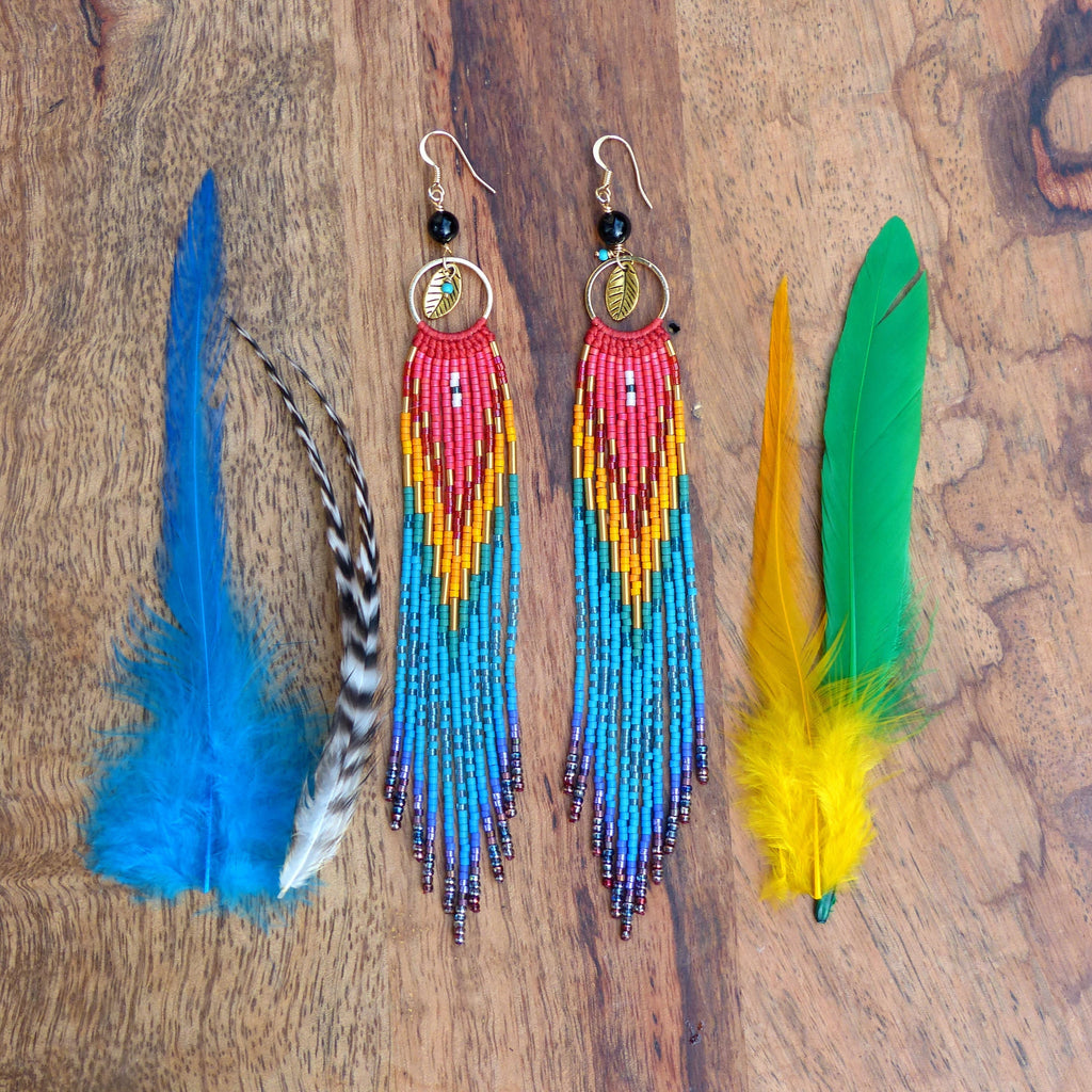 Peut inclure: Une paire de longues boucles d'oreilles en perles colorées avec un charme en feuille d'or. Les boucles d'oreilles sont faites de perles rouges, orange, jaunes, bleues et violettes. Les boucles d'oreilles sont suspendues à une surface en bois.