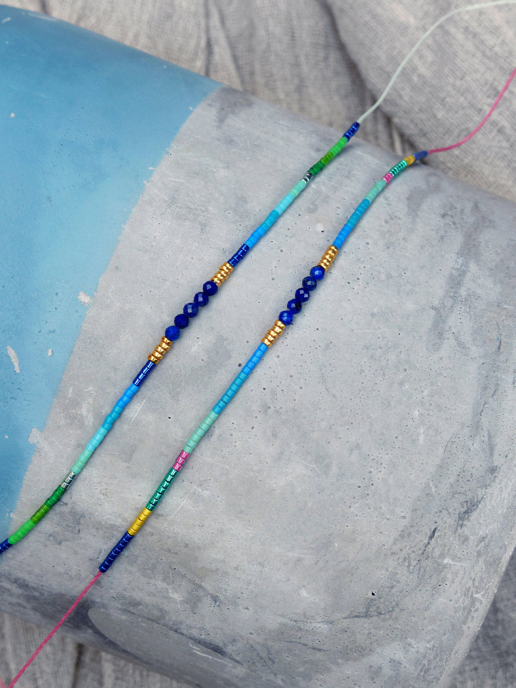 Peut inclure: Deux colliers de perles avec des perles bleues, vertes et dorées. Les colliers sont disposés sur une surface en béton gris.