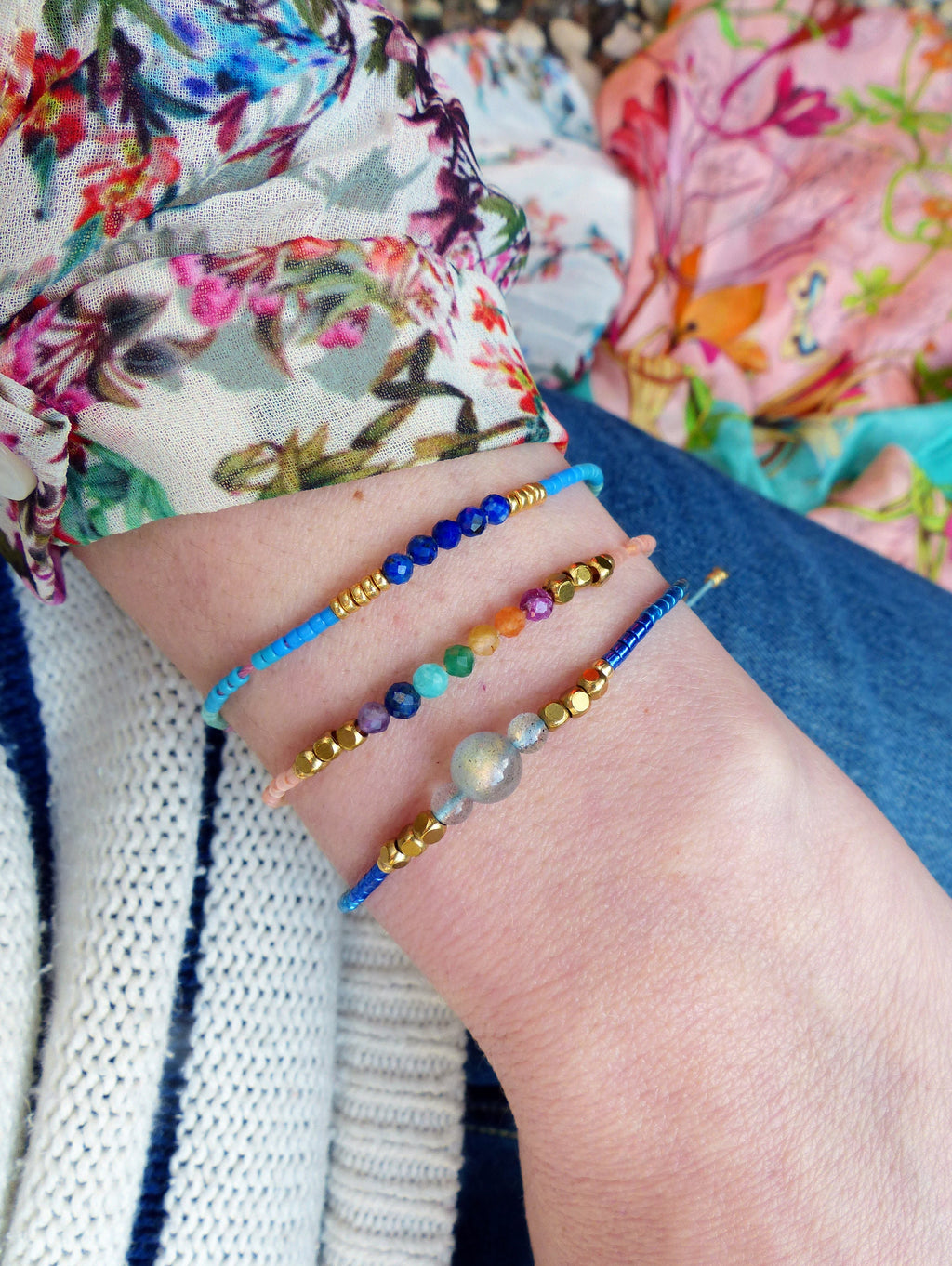 Peut inclure: Trois bracelets en perles sur un poignet. Le bracelet du haut est bleu avec des perles dorées, le bracelet du milieu est orange, rose et vert avec des perles dorées, et le bracelet du bas est bleu avec des perles dorées et une grosse perle irisée.