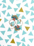 Peut inclure: Deux paires de boucles d'oreilles faites à la main avec des cerceaux dorés et des détails en macramé colorés. Le macramé présente des perles turquoise, roses et dorées. Une boucle d'oreille a une petite perle blanche. Les boucles d'oreilles sont présentées sur une surface blanche avec des motifs de triangles bleus et dorés.