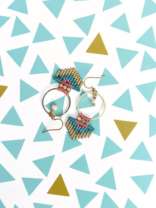Peut inclure: Deux paires de boucles d'oreilles faites à la main avec des cerceaux dorés et des détails en macramé colorés. Le macramé présente des perles turquoise, roses et dorées. Une boucle d'oreille a une petite perle blanche. Les boucles d'oreilles sont présentées sur une surface blanche avec des motifs de triangles bleus et dorés.