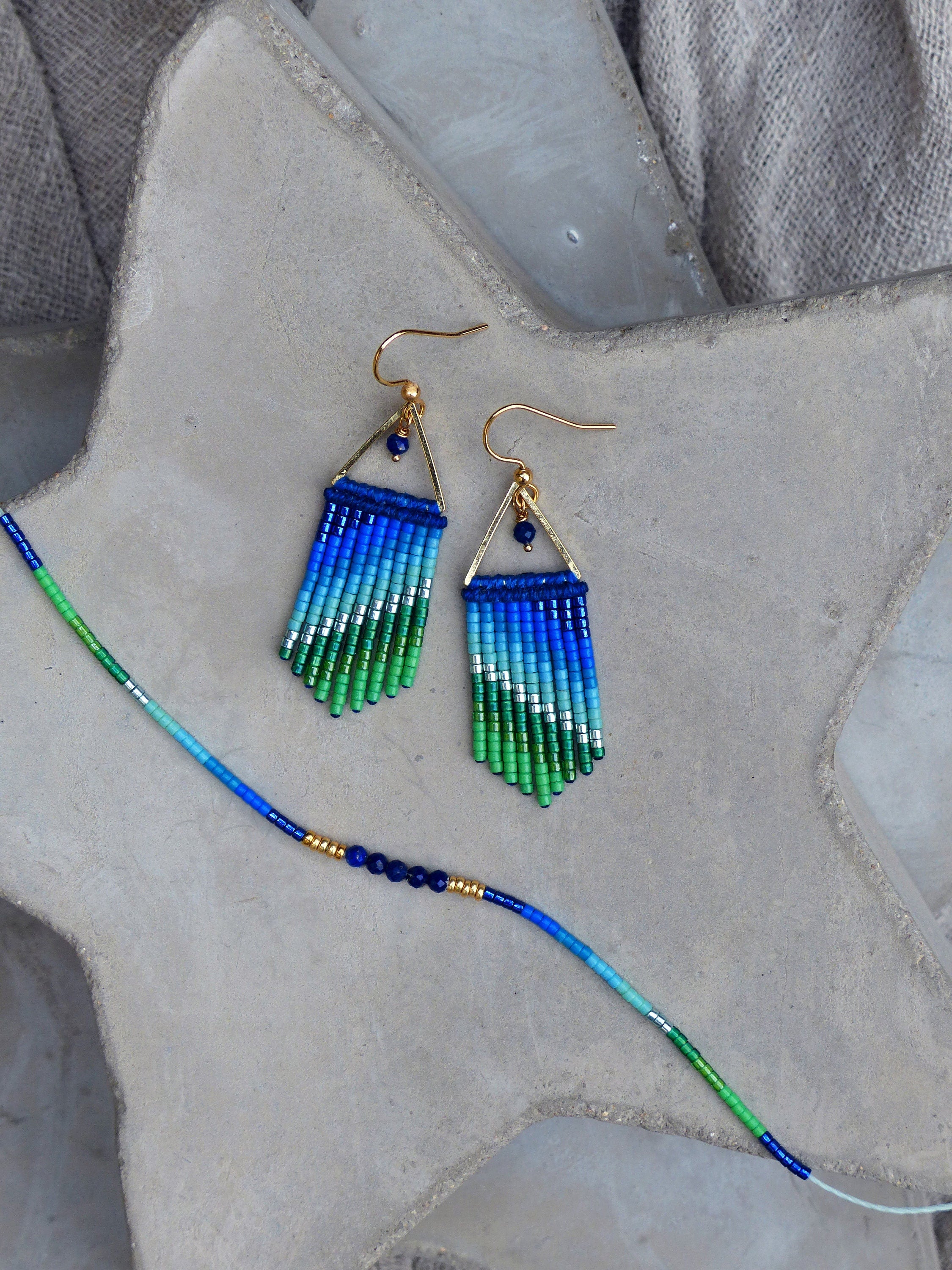 Peut inclure: Une paire de boucles d'oreilles en perles bleues, vertes et argentées avec des sommets triangulaires dorés et des perles de lapis-lazuli. Les boucles d'oreilles sont présentées sur une surface en béton gris avec un collier de perles assorti.