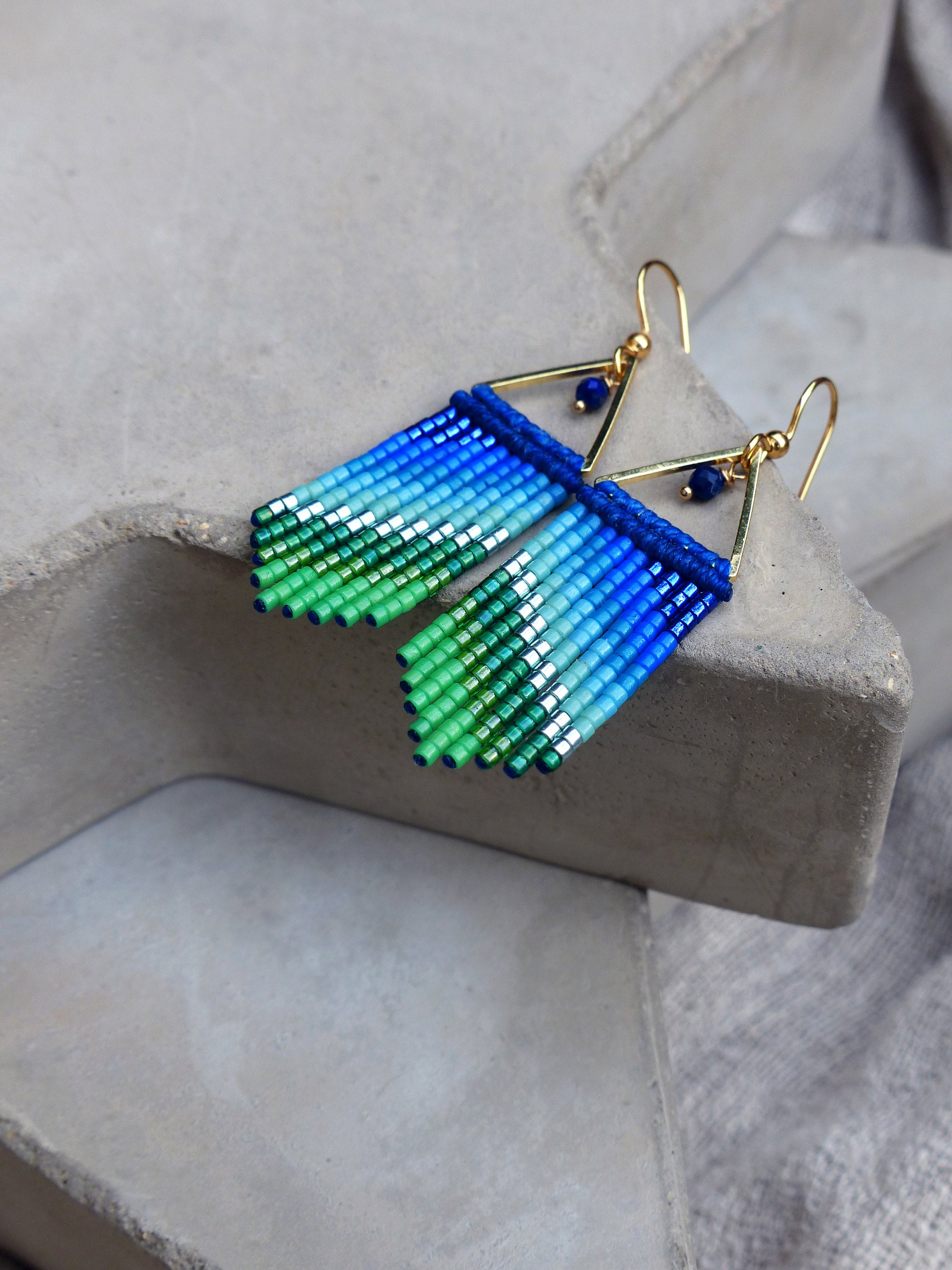 Peut inclure: Une paire de boucles d'oreilles pendantes dorées avec des franges de perles bleues, vertes et argentées. Les boucles d'oreilles ont une forme triangulaire en haut.