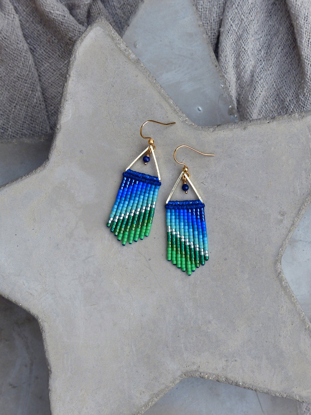 Peut inclure: Une paire de boucles d'oreilles pendantes dorées avec des franges de perles bleues et vertes. Les boucles d'oreilles présentent une petite forme triangulaire avec une pierre précieuse bleue en haut.