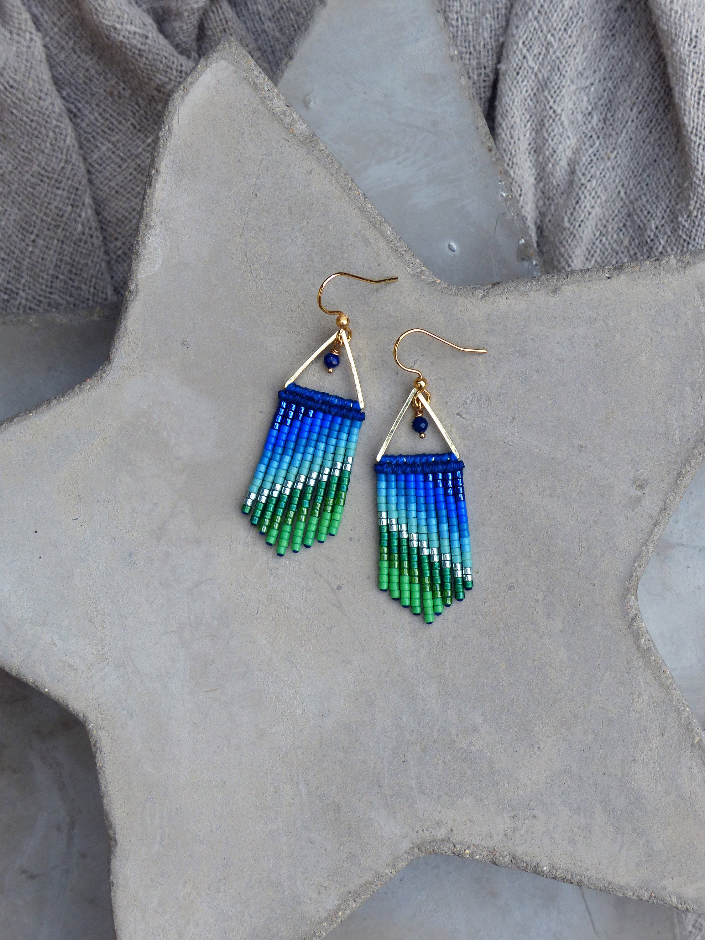 Peut inclure: Une paire de boucles d'oreilles pendantes dorées avec des franges de perles bleues et vertes. Les boucles d'oreilles présentent une petite forme triangulaire avec une pierre précieuse bleue en haut.