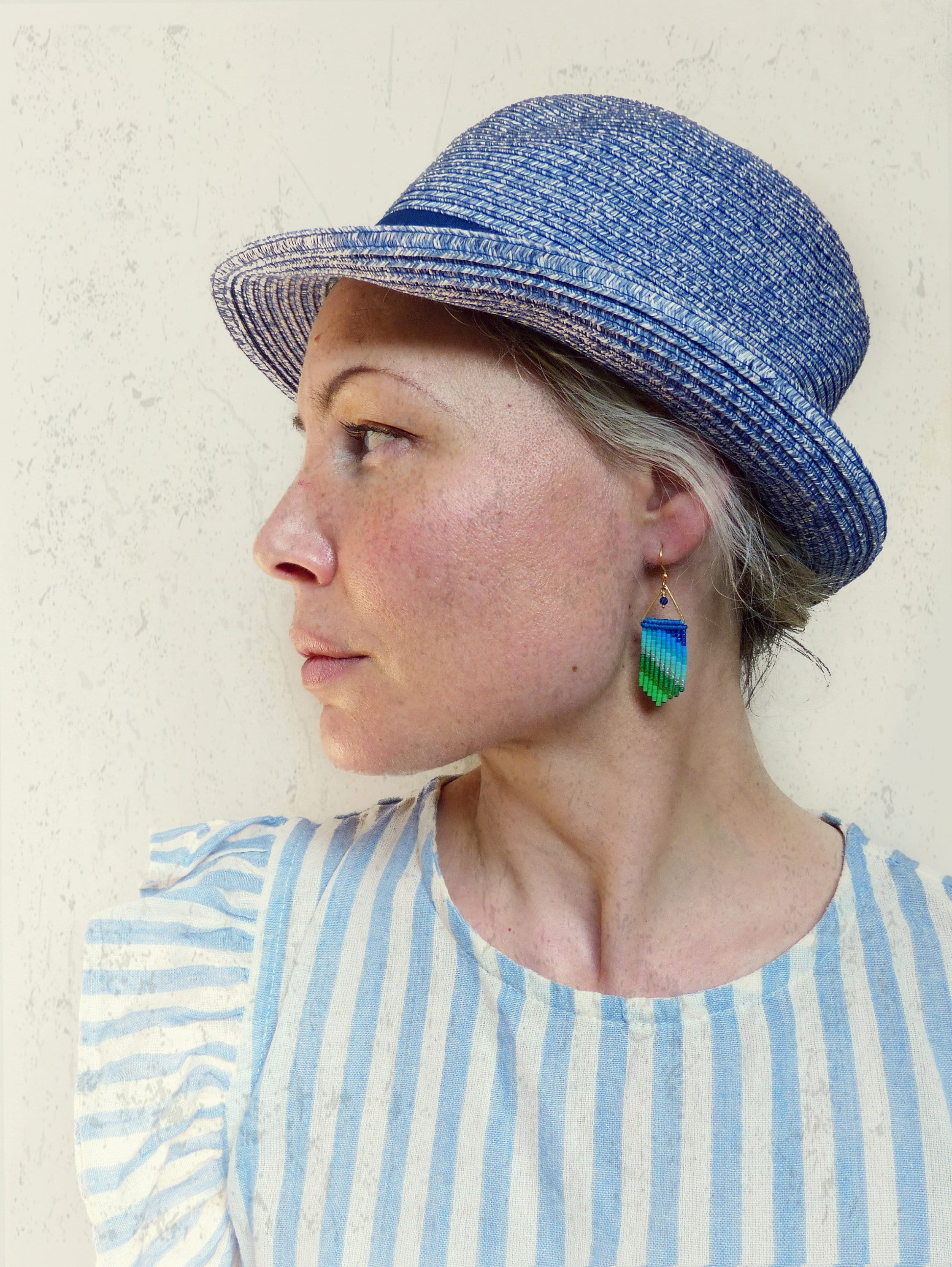 Peut inclure: Une femme portant une robe à rayures bleues et blanches et un chapeau de paille bleu avec un ruban noir. Elle porte une paire de boucles d'oreilles pendantes vertes, bleues et violettes.
