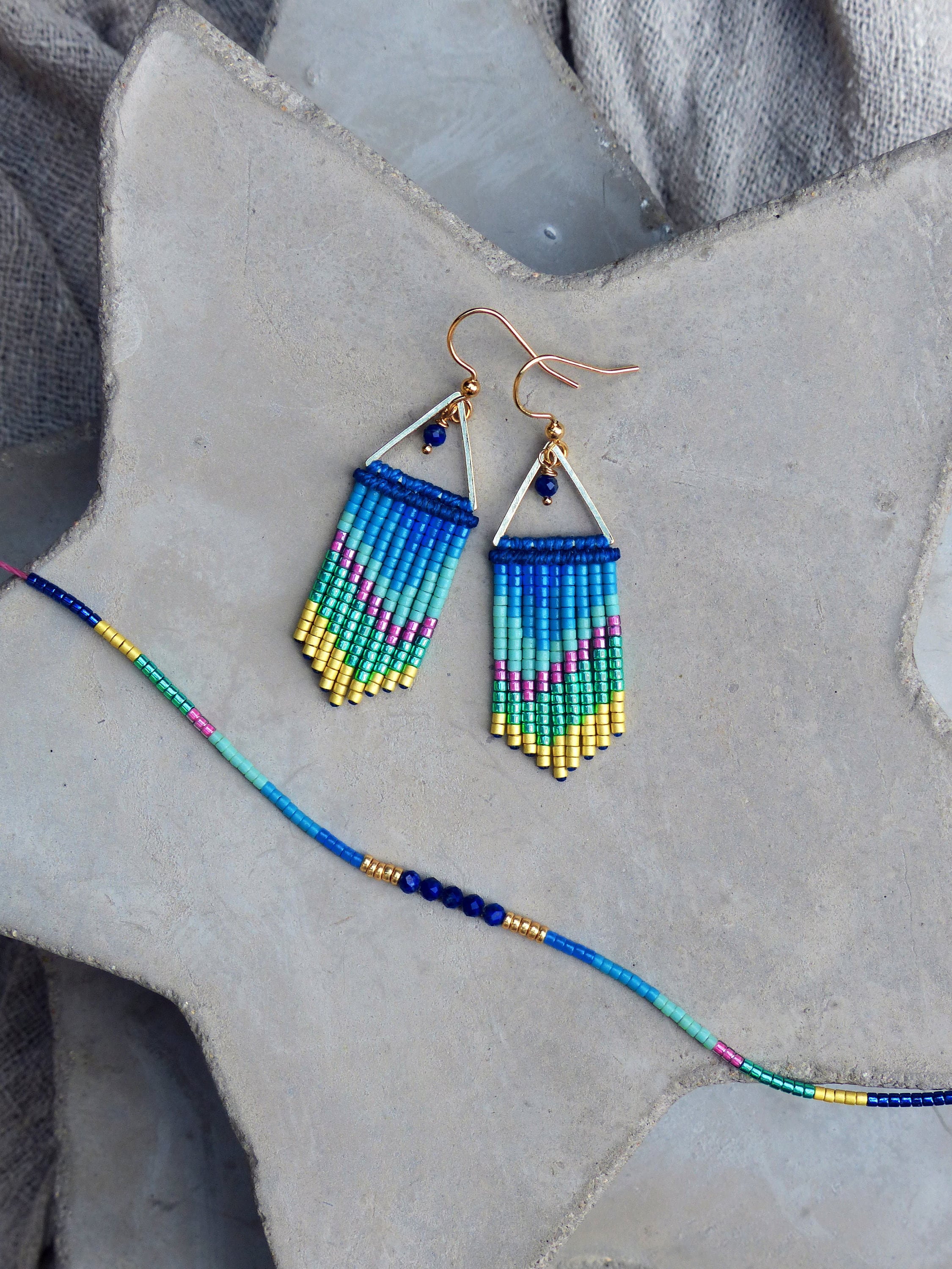 Peut inclure: Une paire de boucles d'oreilles en perles bleues, vertes, roses et dorées de forme triangulaire avec un crochet doré. Les boucles d'oreilles sont sur une surface grise avec un collier de perles bleues, vertes et dorées.