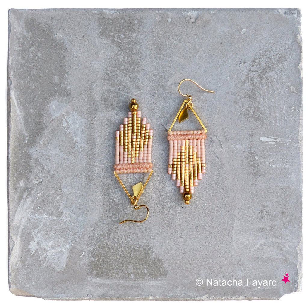Peut inclure: Une paire de boucles d'oreilles en perles dorées et rose poudré avec un design géométrique triangulaire. Les boucles d'oreilles sont suspendues à une surface grise.