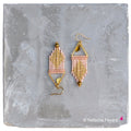 Peut inclure: Une paire de boucles d'oreilles en perles dorées et rose poudré avec un design géométrique triangulaire. Les boucles d'oreilles sont suspendues à une surface grise.