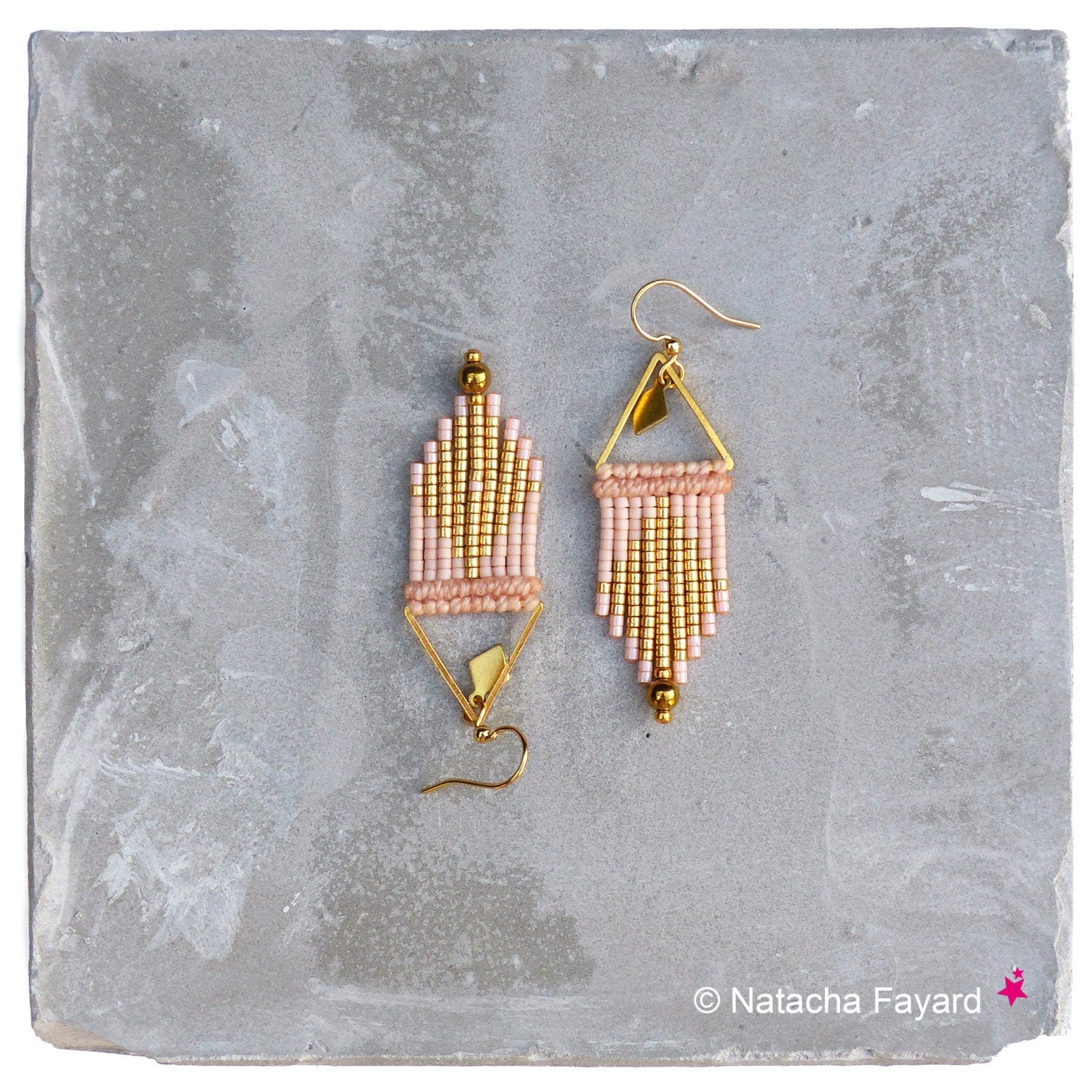 Peut inclure: Une paire de boucles d'oreilles en perles dorées et rose poudré avec un design géométrique triangulaire. Les boucles d'oreilles sont suspendues à une surface grise.
