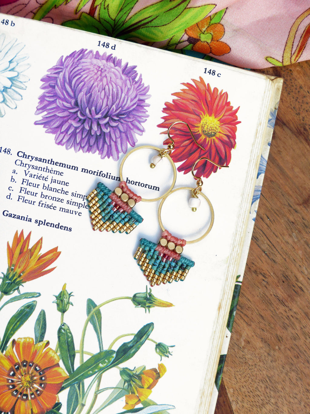 Peut inclure: Boucles d'oreilles créoles dorées avec des détails en macramé turquoise, or et rose. Les boucles d'oreilles sont présentées sur une illustration botanique vintage de fleurs. Elles ont une petite perle blanche et un crochet doré.