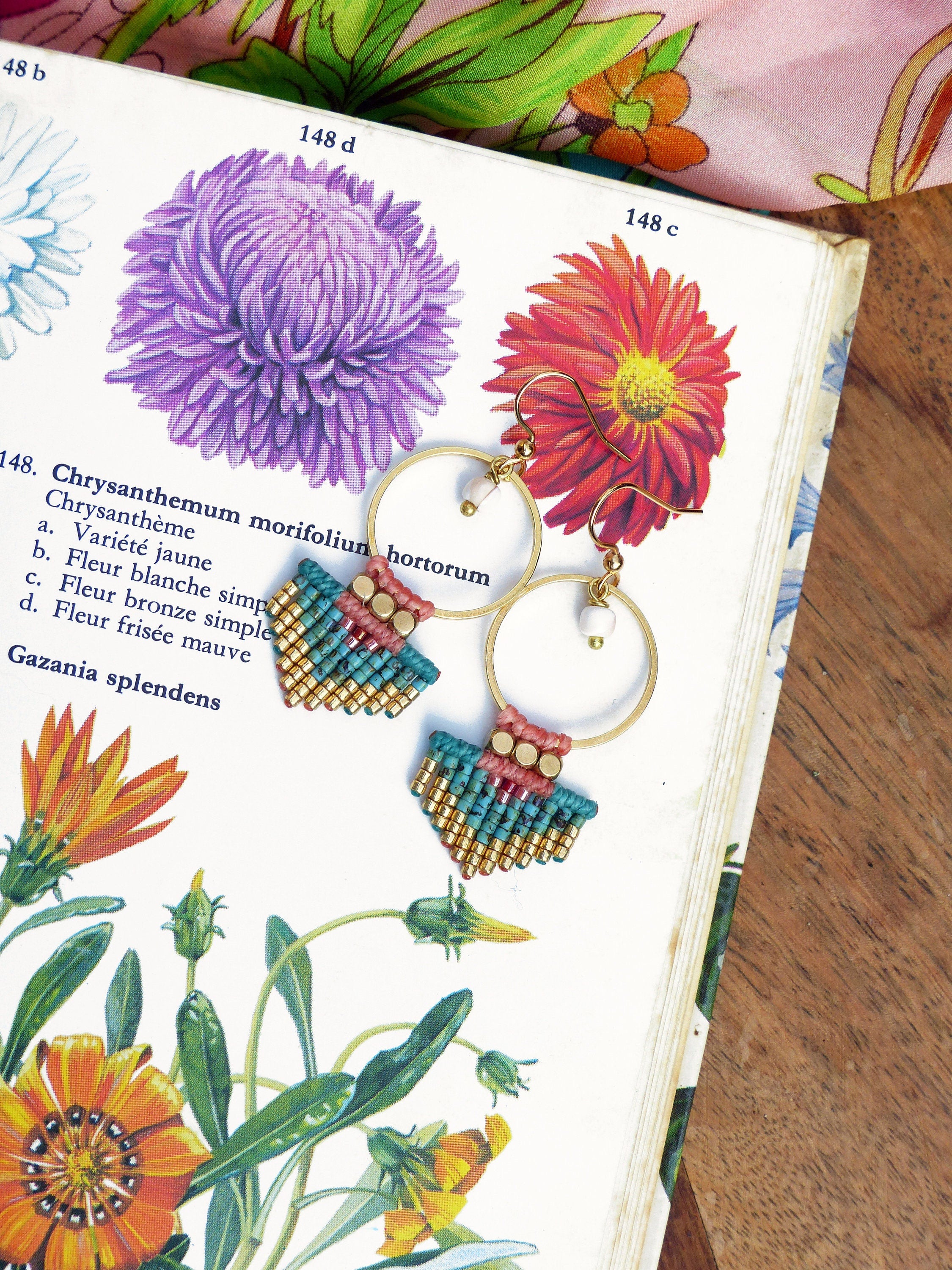 Peut inclure: Boucles d'oreilles créoles dorées avec des détails en macramé turquoise, or et rose. Les boucles d'oreilles sont présentées sur une illustration botanique vintage de fleurs. Elles ont une petite perle blanche et un crochet doré.