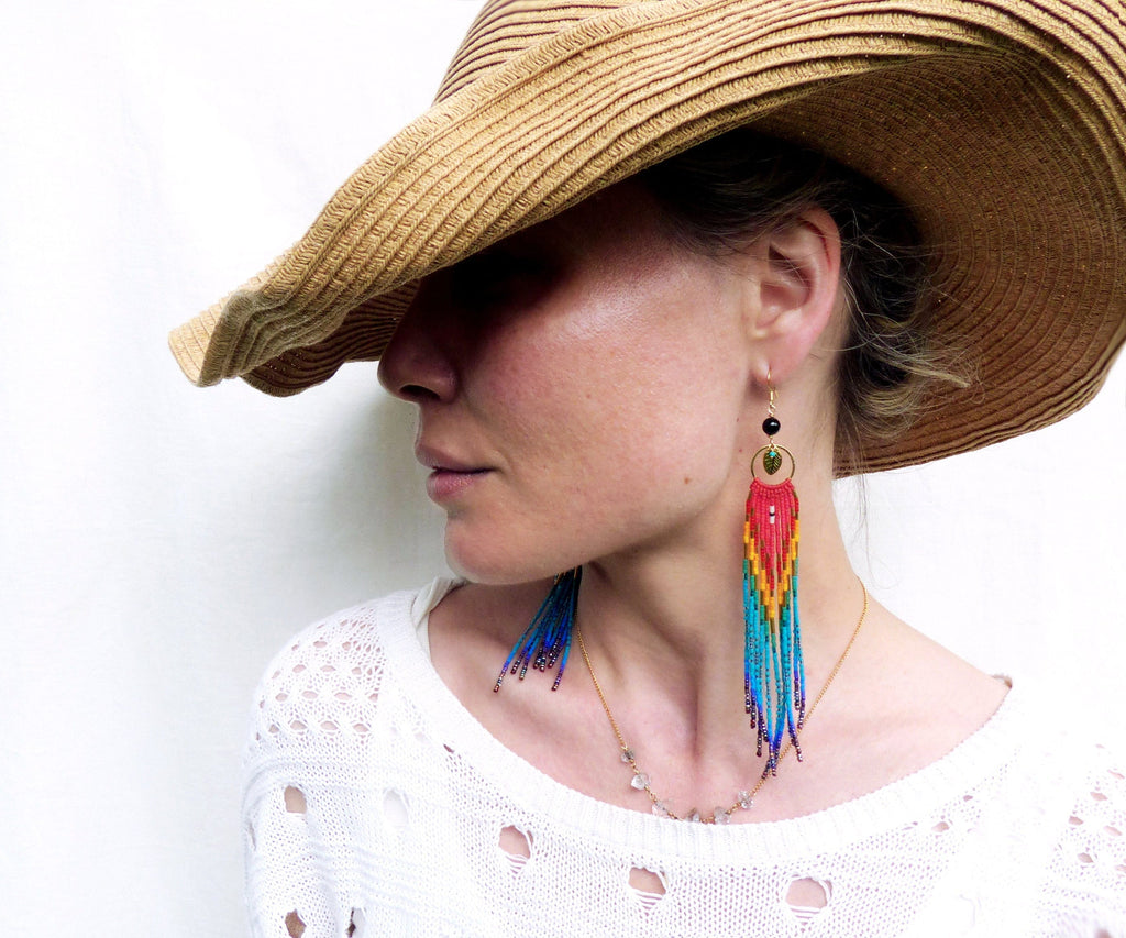 Peut inclure: Une femme portant un chapeau de paille et un pull blanc avec un collier de perles. Elle porte de longues boucles d'oreilles en perles colorées avec un accent de pierre verte.