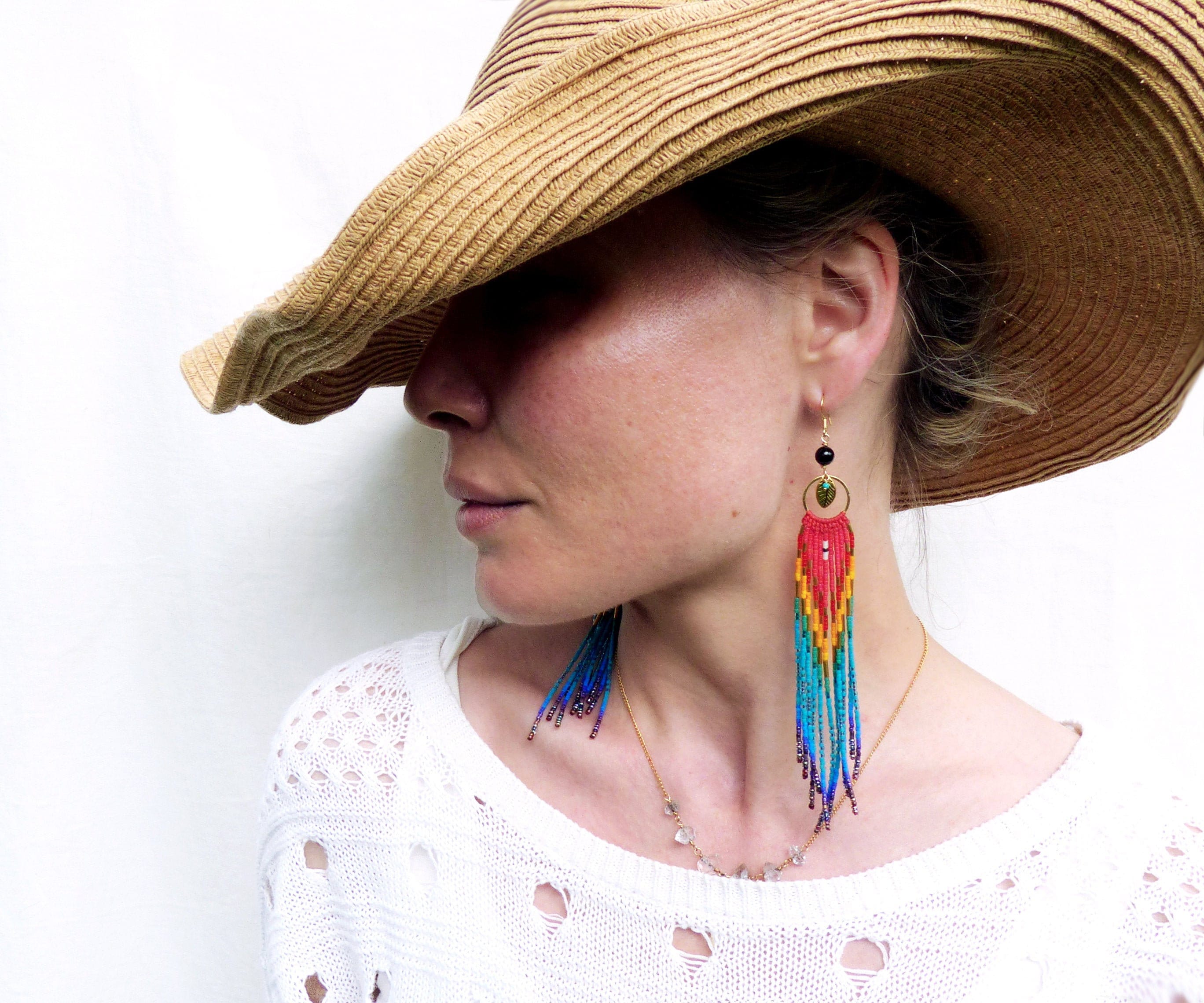 Peut inclure: Une femme portant un chapeau de paille et un pull blanc avec un collier de perles. Elle porte de longues boucles d'oreilles en perles colorées avec un accent de pierre verte.