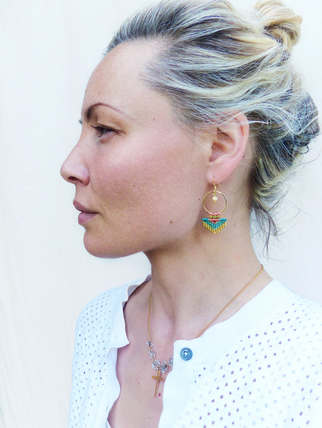 Peut inclure: Boucles d'oreilles dorées avec une frange de perles turquoise, jaunes et roses. Un collier doré avec un pendentif en forme de croix et un cardigan blanc sont également visibles.