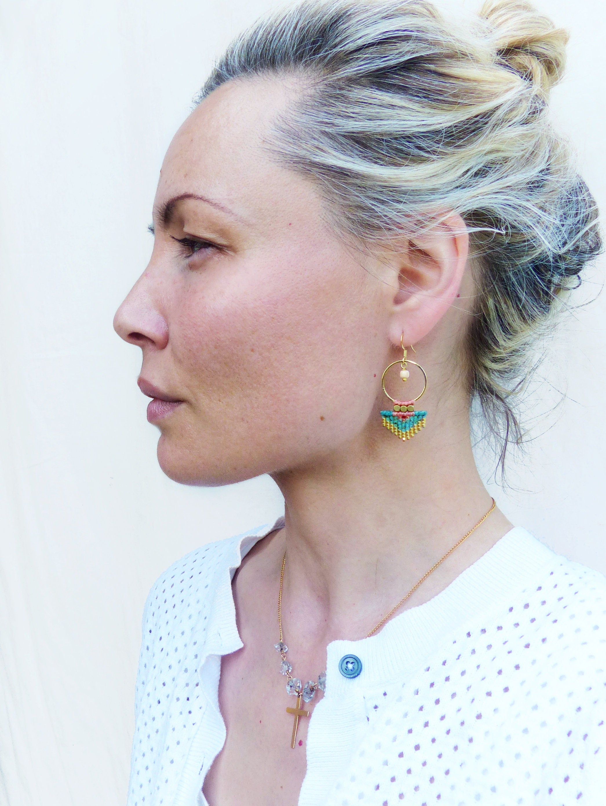 Peut inclure: Boucles d'oreilles dorées avec une frange de perles turquoise, jaunes et roses. Un collier doré avec un pendentif en forme de croix et un cardigan blanc sont également visibles.