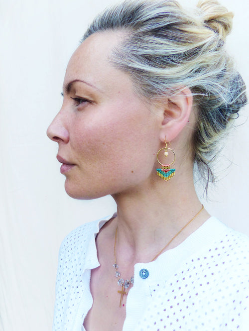 Peut inclure: Boucles d'oreilles dorées avec une frange de perles turquoise, jaunes et roses. Un collier doré avec un pendentif en forme de croix et un cardigan blanc sont également visibles.