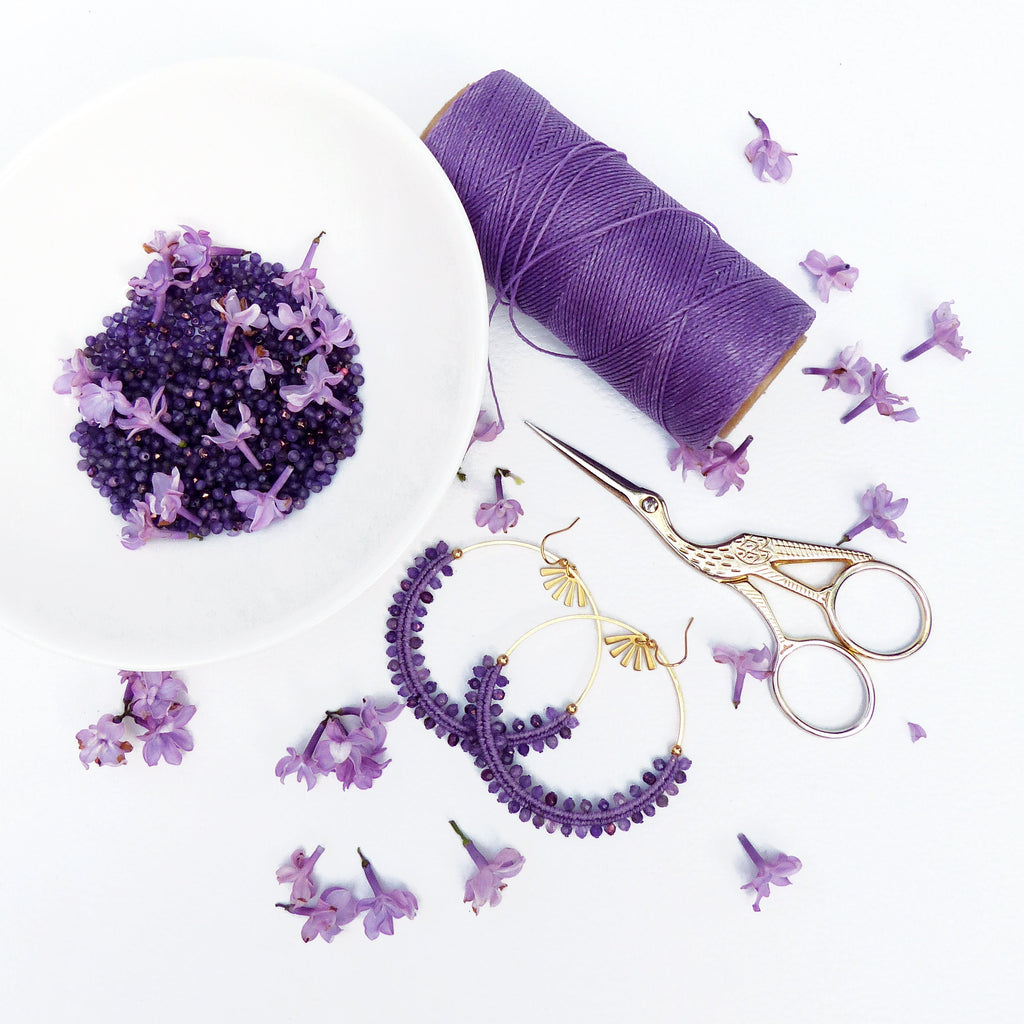 Peut inclure: Une image de nature morte présentant des fournitures d'artisanat et des bijoux. Un bol blanc contient de petites perles violettes et des fleurs de lilas. Une bobine de fil violet, des ciseaux dorés et deux paires de boucles d'oreilles créoles perlées violettes sont également présents.