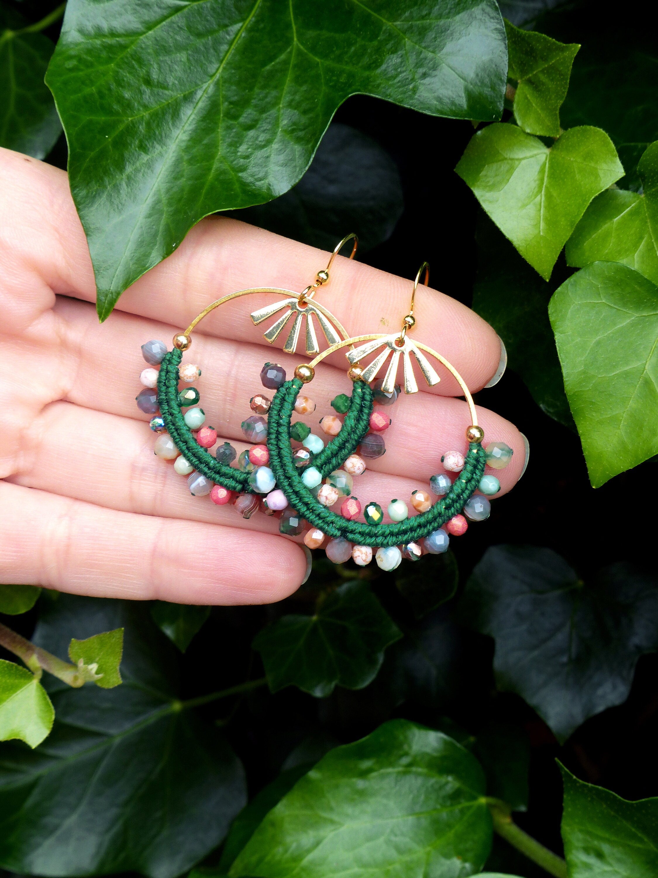 Peut inclure: Une paire de boucles d'oreilles créoles vertes et dorées avec des perles colorées. Les boucles d'oreilles sont faites de fil vert et ont un anneau doré avec un motif en forme d'éventail.